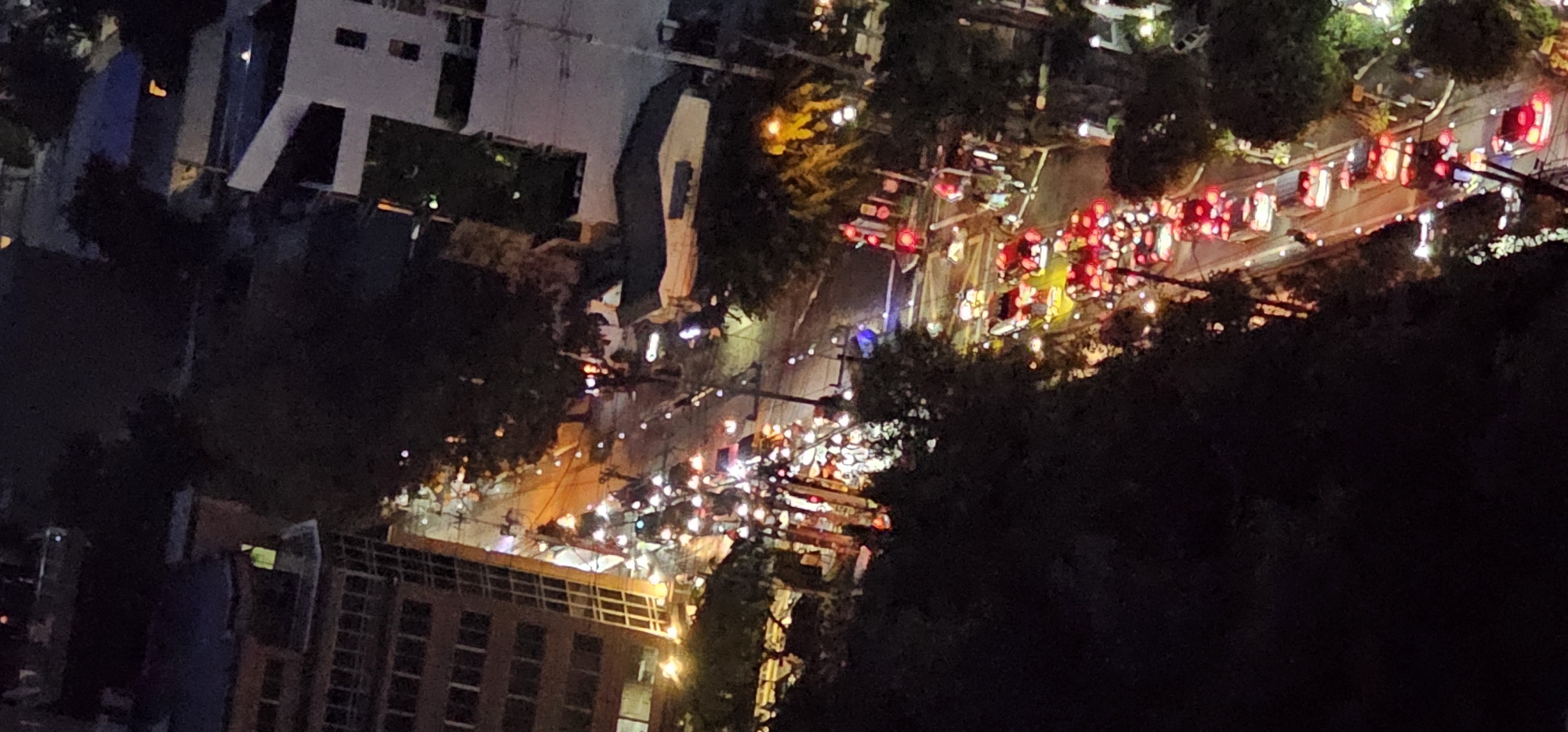 The image captures a nighttime aerial view of a traffic jam on a city street, likely in Makati, Philippines. The scene is dominated by the red taillights of numerous vehicles, indicating slow or stopped movement. Several buildings are visible alongside the street, some illuminated, suggesting commercial or residential areas. Trees line parts of the road, their dark foliage contrasting with the artificial lights. The overall impression is one of urban congestion under artificial illumination.