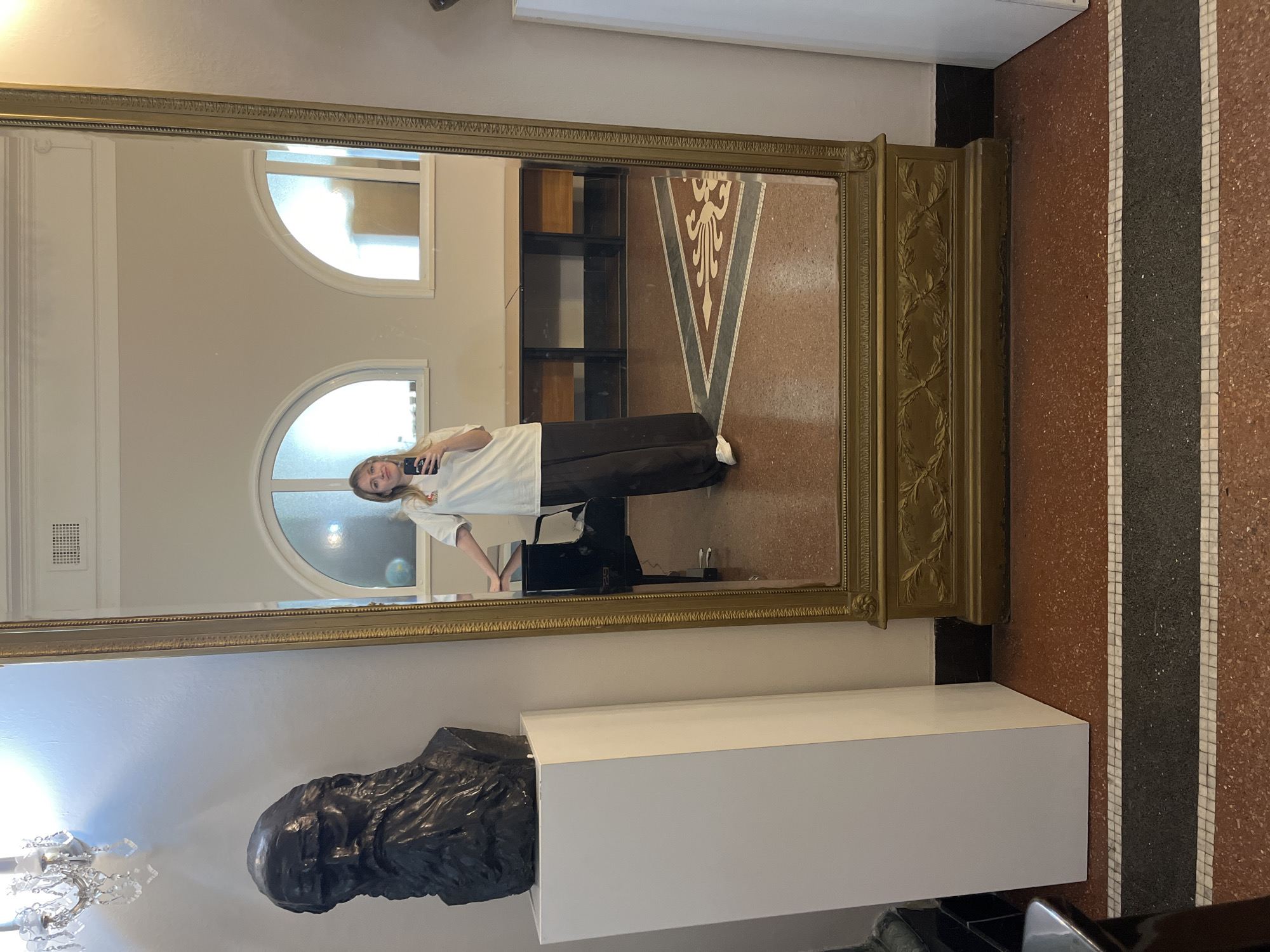 A young woman stands in front of a large, ornate, gold-framed mirror, taking a selfie with her phone. She wears a white t-shirt and black pants. The reflection shows part of a room with arched windows, a dark bookshelf, and a patterned floor. In the foreground, outside the mirror, a dark bust sits on a white pedestal. The floor visible outside the mirror has a red, black, and white striped pattern. The location is Belgrade, Serbia.