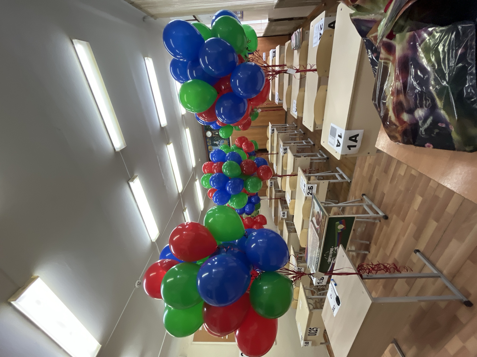 Here's a forensic description of the image:
The image appears to be taken inside a classroom or similar educational setting. Rows of student desks are visible, each with a small numbered label attached. Some desks have additional printed materials on them.
Floating above the desks are clusters of red, green, and blue balloons. The balloons are tethered to the desks.
The room is illuminated by fluorescent lights mounted on the ceiling. The floor appears to be made of wood or laminate.
The OCR data includes terms like "ПАРТА ГЕРОЯ" (Parta Geroya), suggesting a commemorative or memorial aspect, and labels "1A", "1B", "2A", etc., likely referencing seat assignments. The OCR also detects a brand name "Daci", possibly associated with the materials on the desks.
Based on the "ПАРТА ГЕРОЯ" (Parta Geroya) reference in the OCR and a quick internet search, the classroom's location is potentially in Kaspiysk, Russia. The phrase translates to "Hero's Desk" and is part of a memorial project within Russian schools.