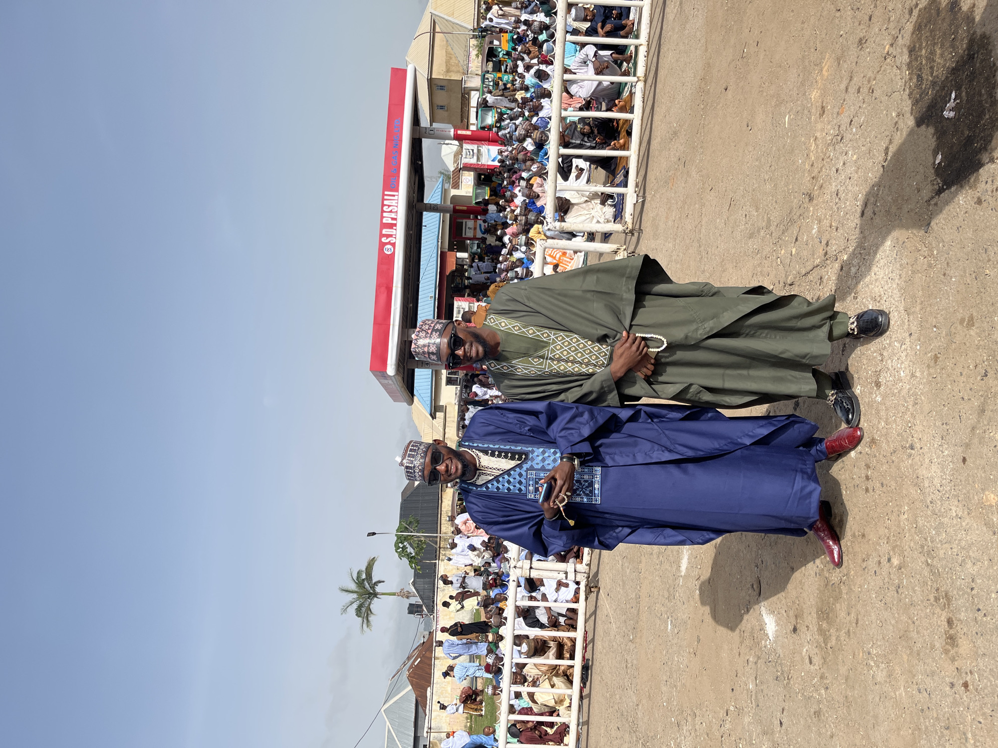 Two men stand on a concrete surface, facing the camera. The man in front wears a blue robe and red shoes, and holds what appears to be a phone and prayer beads. The man behind him is dressed in a green robe and black shoes, also holding prayer beads. Both men wear hats with similar patterns. Behind the men is a white metal fence, and behind the fence, a large crowd of people is seated. Further behind the crowd, a red-and-white structure is visible, with the text "S.D. PASALI" in white letters on a red background, likely indicating a business or station. A palm tree is visible to the left of the men. The location is determined to be in Jos, Nigeria.