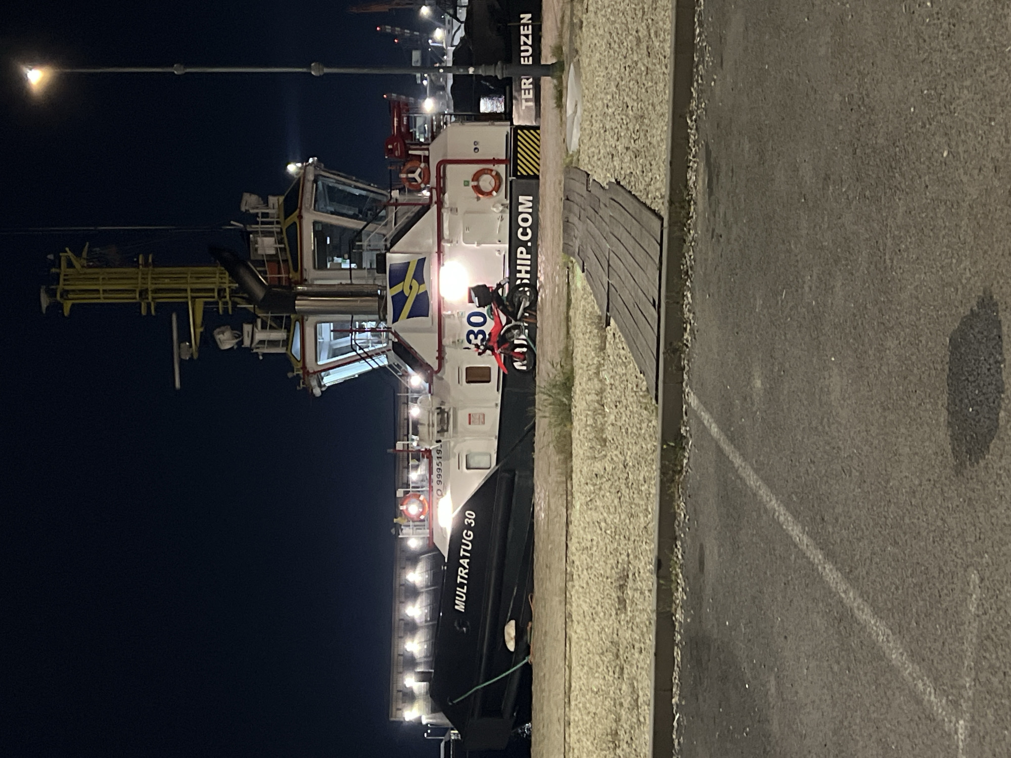 The image depicts a port scene at night, likely in Burgas, Bulgaria, given the tugboat's name. A tugboat named "MULTRATUG 30" is docked along a concrete quay. A red and white motorcycle is parked alongside the tugboat. To the right of the boat is a walkway covered in gray wood planks. The surface of the quay is dark asphalt with a white line marking a lane or border. A lamppost illuminates the foreground. The overall setting is industrial, with a dark night sky.