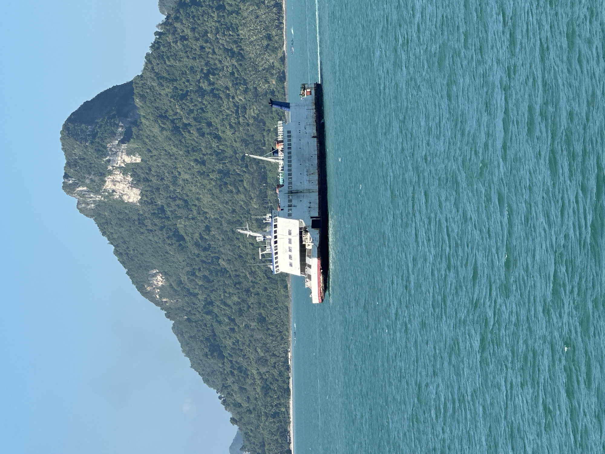 A ferry boat navigates the turquoise waters of the Gulf of Thailand off the coast of Don Sak, Thailand. The ferry, a long, white vessel with blue accents, is positioned centrally in the frame and appears to be moving from left to right. In the background, a lush, green mountainous island dominates the horizon, its slopes covered in dense vegetation. A narrow strip of sandy beach is visible at the base of the island, separating the water from the forest. The sky above is a clear, bright blue. The water is choppy with visible ripples and small waves, indicating a breezy day. The scene conveys a sense of travel and the natural beauty of the coastal region.