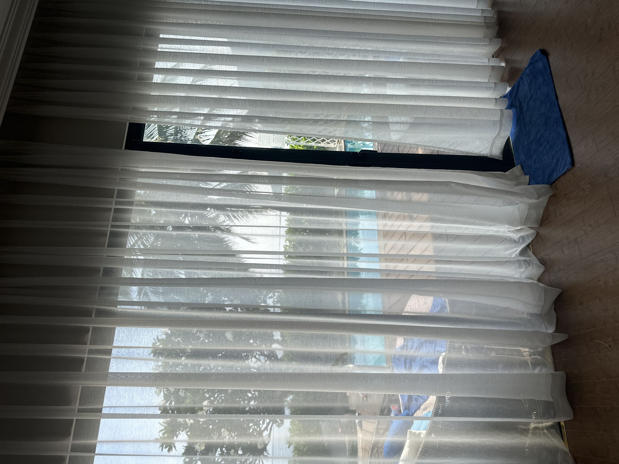 A room in the city of Unknown, Unknown, presents a sunlit view through white, sheer curtains covering a window. Outside, a scene featuring palm trees, sand, and water is partially visible. A folded blue towel lies on the wooden floor next to the curtain.