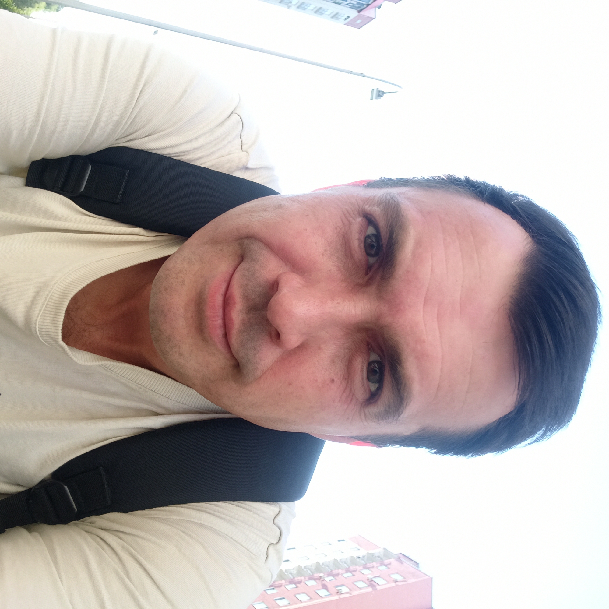 A man is taking a selfie outdoors. He is wearing a light-colored t-shirt and a black backpack. His face is turned towards the camera, and he is smiling slightly. In the background, there is a bright sky, a street lamp, and buildings. The buildings appear to be residential and are located in Nizhny Novgorod, Russia. One building is pink and white with multiple windows, and another building is white with red trim. The overall scene suggests a casual, daytime setting in an urban environment.