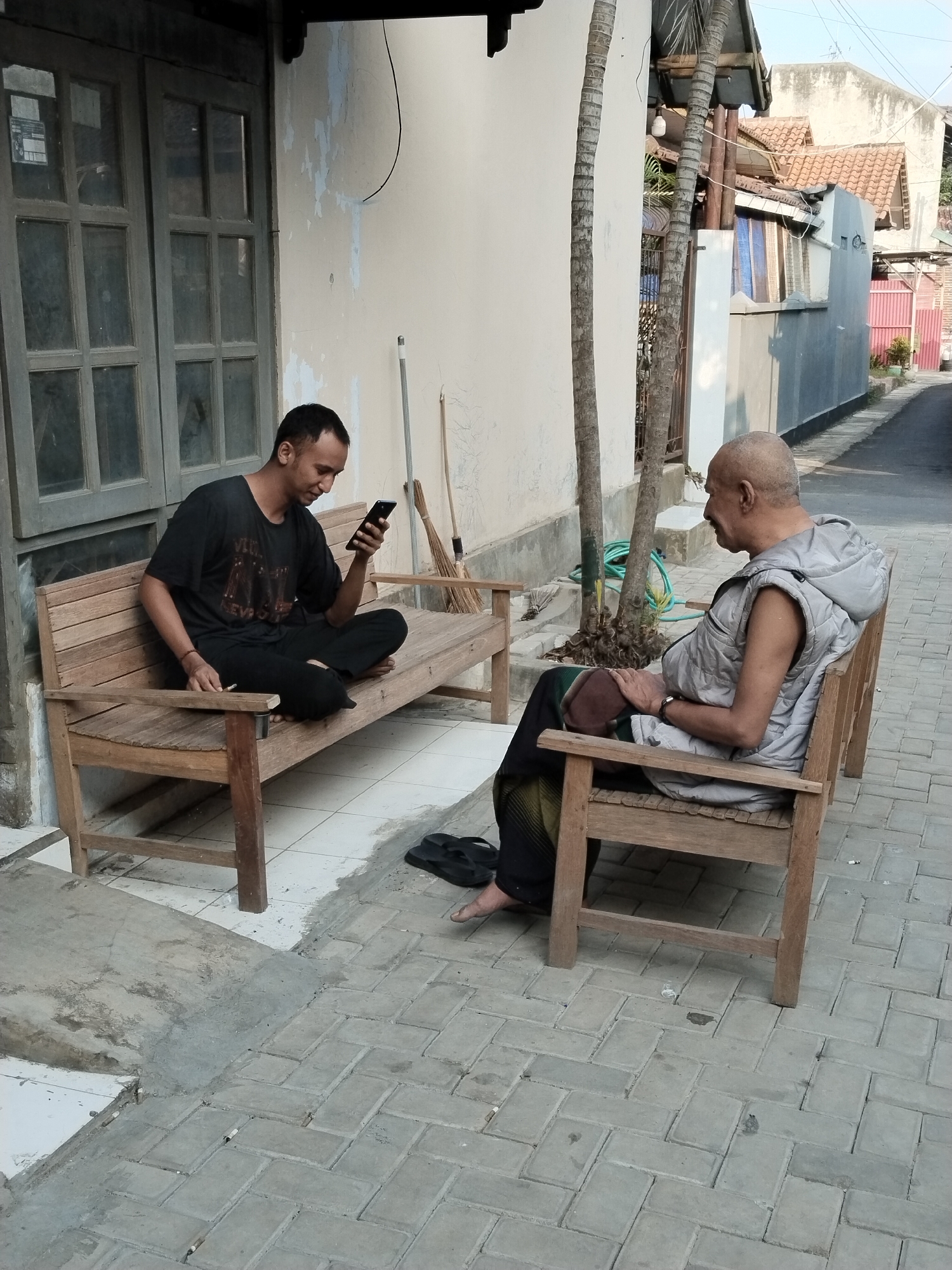 Two men are seated on wooden benches outside a building, likely in a residential area of Kecamatan Pati, Indonesia. The man on the left, wearing a black t-shirt and black pants, is sitting cross-legged and looking at his smartphone. He appears to be holding a cigarette in his right hand. The man on the right, an older gentleman with a shaved head and wearing a light gray vest over dark pants, is seated facing away from the camera, looking to his left. He has his hands clasped in his lap. Behind the men, a textured wall shows signs of wear, and a wooden door with glass panes is visible. The ground is paved with interlocking concrete bricks. A tree with a rough, segmented trunk stands between the two men, and further down the paved area, houses with red-tiled roofs are visible, suggesting an urban or semi-urban environment. The lighting suggests it is daytime, with clear skies.