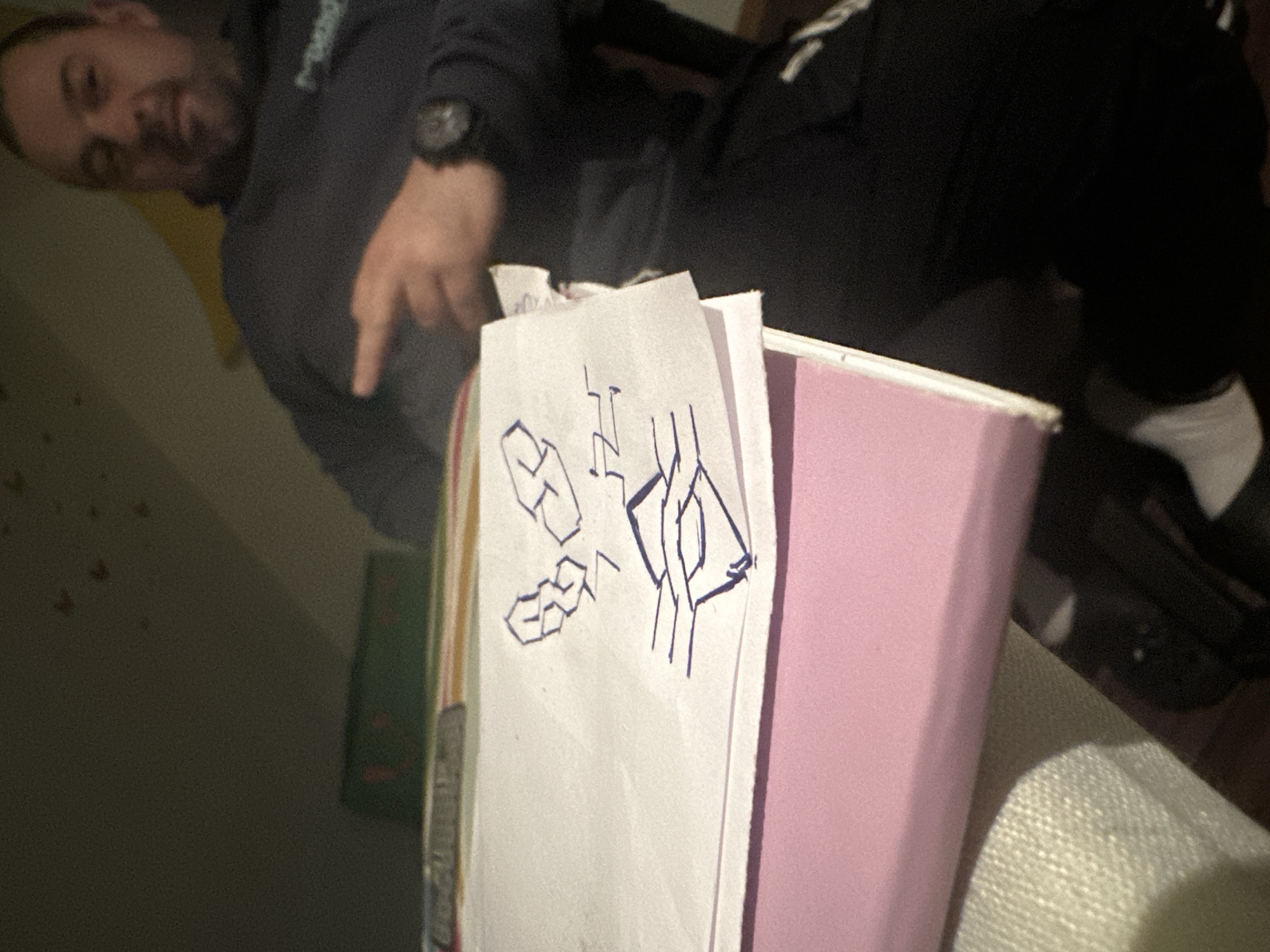 A man is seated, leaning over a desk or table. He is wearing a dark-colored sweatshirt and dark pants with white stripes. He is pointing at a piece of white paper with blue pen drawings. The paper is on top of a pink folder. The background includes a wall with possible shadow details. The location is not evident from the image.