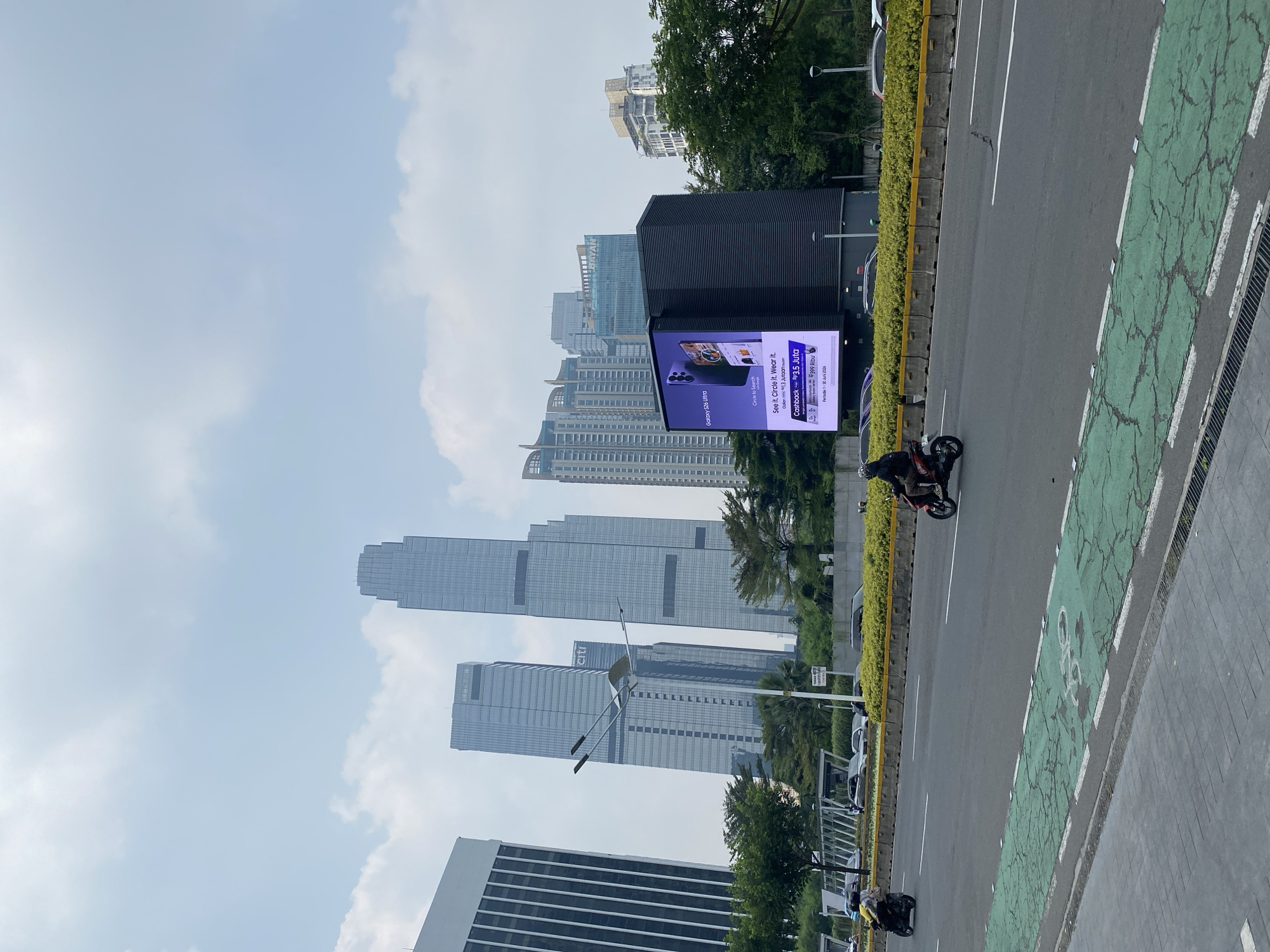 This is an eye-level, full shot of a street scene in Jakarta, Indonesia, on a cloudy day. In the foreground, a paved sidewalk runs along the bottom edge of the frame. To its right is a street with a green bike lane marked with white lines and a bicycle symbol. Two motorcycles are visible on the road: one in the left lane, moving away from the camera, and another in the right lane, also moving away from the camera.
Across the street, there is a median strip covered in yellow-green shrubbery, bordered by concrete barriers. Beyond the median, a line of cars is visible. Further in the background, a large, modern building with a black facade stands prominently, displaying a large digital billboard. The billboard shows an advertisement for the "Galaxy S26 Ultra" phone, featuring an image of the phone and promotional text about "Cashback" and "3.5 Juta."
Behind the billboard building, a cluster of tall, modern skyscrapers dominates the skyline. Some of these buildings have glass facades, reflecting the cloudy sky. One skyscraper has the "Citi" logo visible near its top. Trees and other greenery are interspersed between the buildings, softening the urban landscape. The overall scene depicts a busy urban environment with a mix of commercial buildings, traffic, and advertisements.