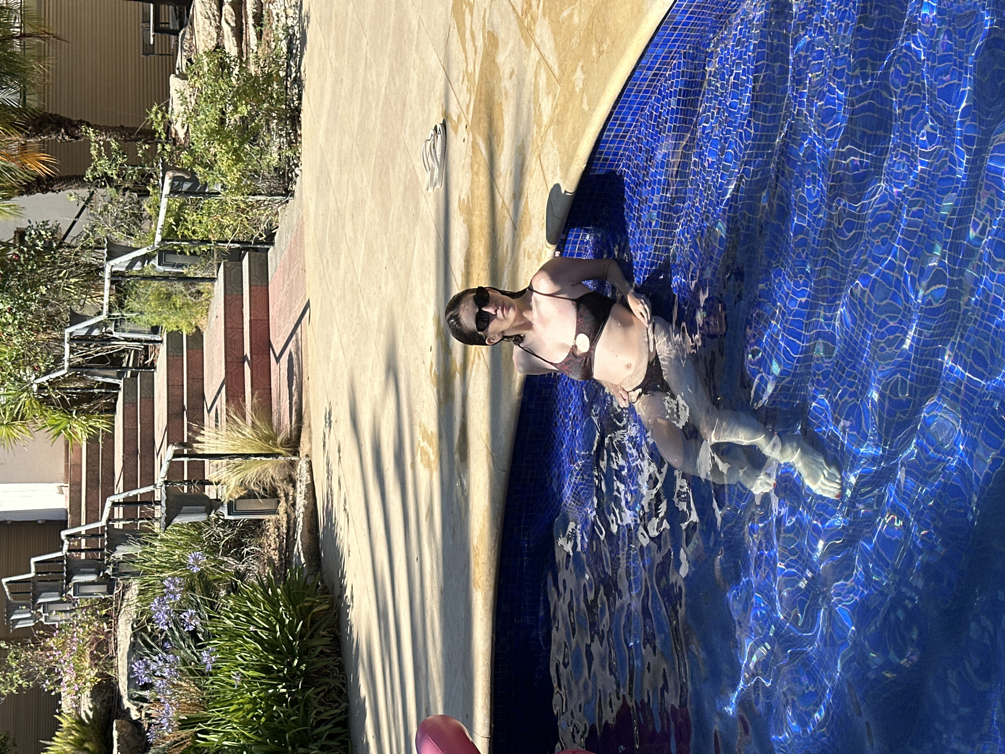 A young woman with dark hair and sunglasses is relaxing in a swimming pool. She is wearing a dark-colored bikini. The pool is tiled with blue mosaic tiles. In the background, there is a stone patio with shadows from trees. Behind the patio, there is a set of outdoor stairs with railings and potted plants. The location is identified as Savyon, Israel.