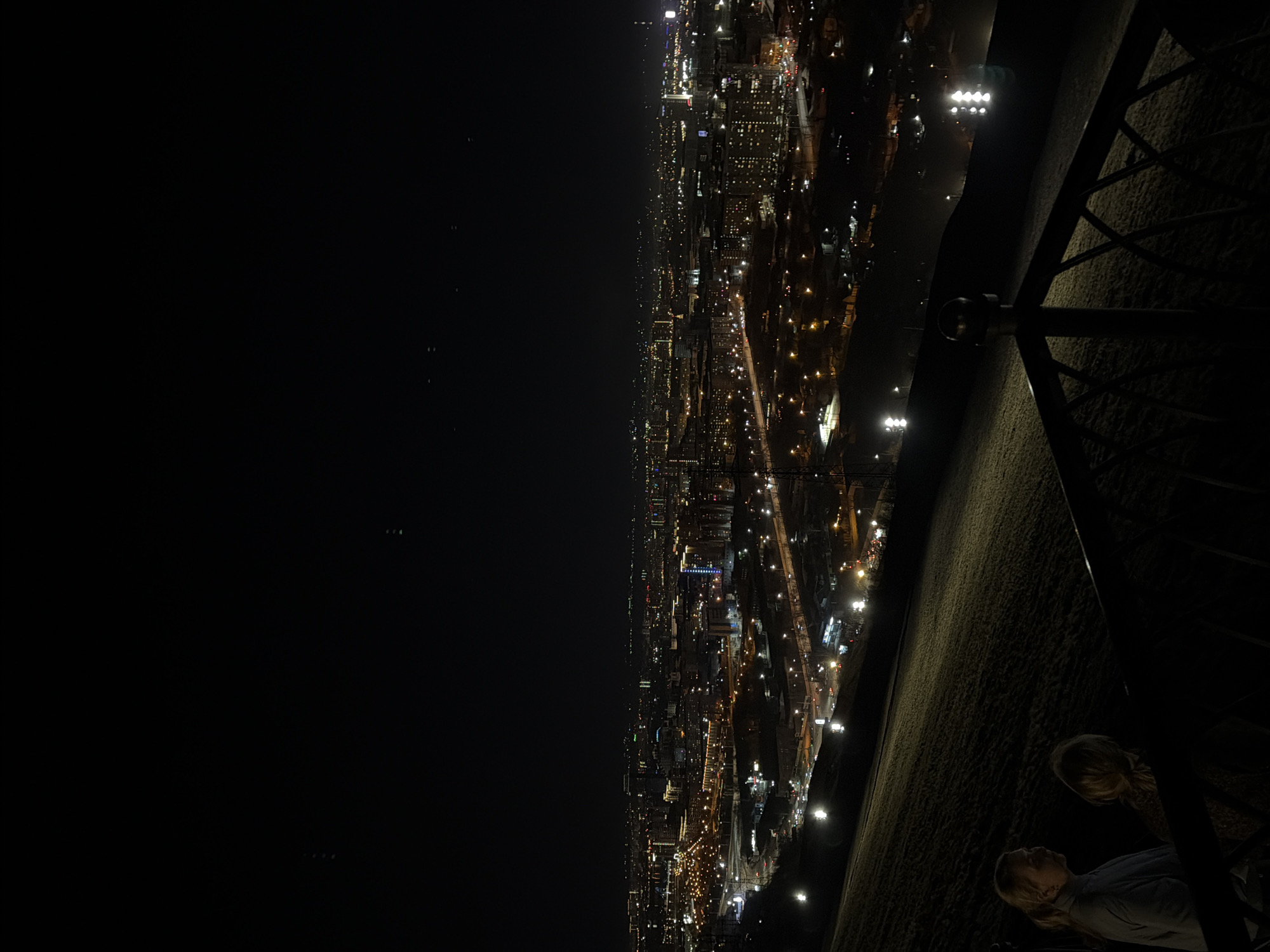 At night, a long, dark, horizontally oriented image shows a city view from a height, with lights scattered across a built environment that stretches far into the distance. A railing runs along the top right of the frame, and two people are visible in the bottom right corner, one looking up towards the city. The scene is dimly lit, with the city lights providing the primary illumination.
The image likely depicts Krasnoyarsk, Russia, because the city is known for its views from high vantage points.