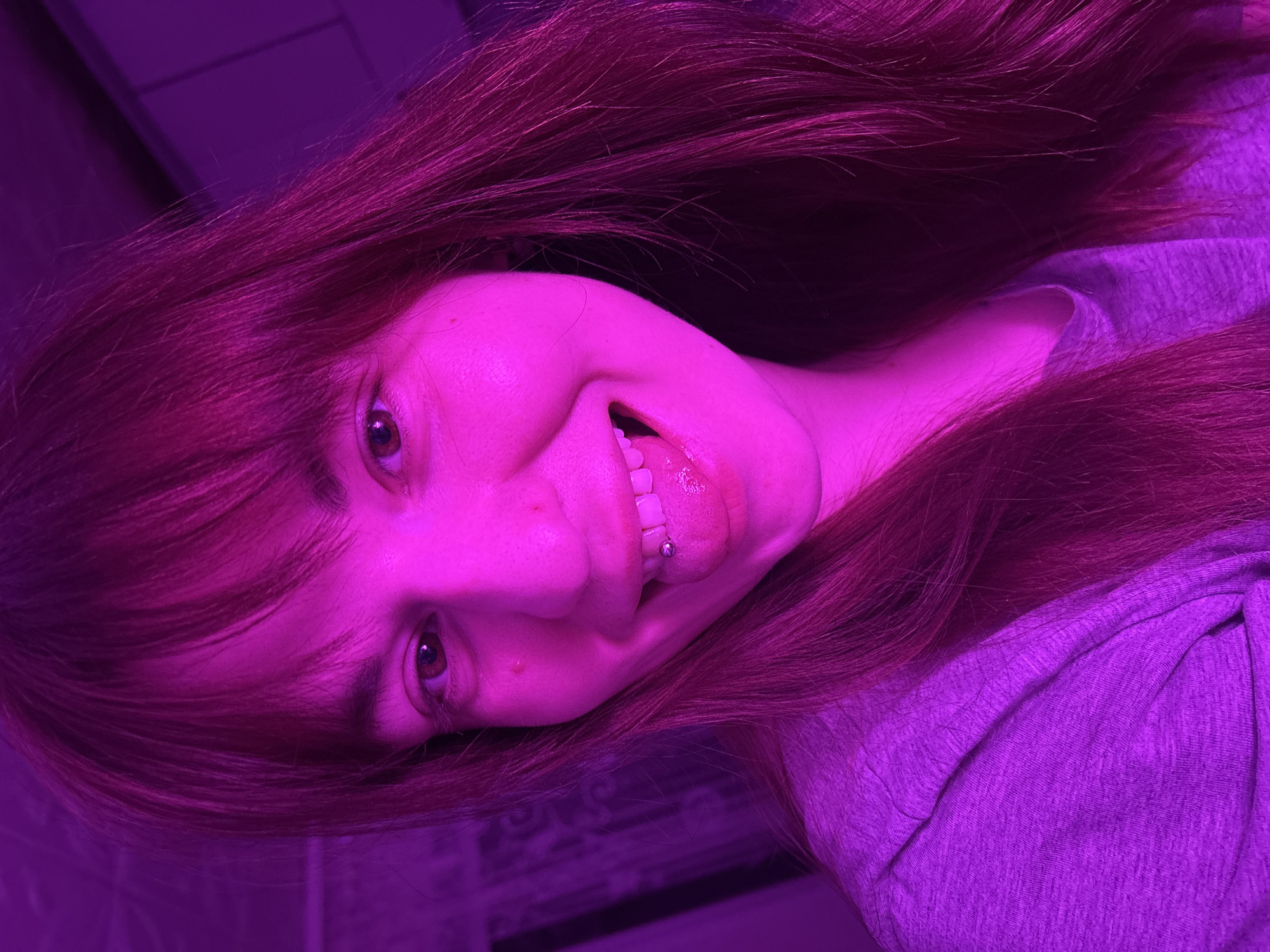 A close-up, angled portrait captures a person, likely a young woman, illuminated by strong purple and magenta lighting. Her long, dark reddish-brown hair cascades around her shoulders, appearing vibrantly magenta under the artificial light. She wears a grey t-shirt, which also takes on a purple hue.
The subject is smiling broadly, her mouth open to reveal white teeth and a tongue piercing. Her brown eyes look directly at the viewer. The overall expression is playful and engaged. The background is dark and indistinct, suggesting an indoor setting. The intense purple lighting dominates the scene, casting a deep color over her skin, hair, and clothing, creating a distinct and dramatic ambiance. No specific location cues are visible within the image to suggest the provided context of "Фоки, Russia." There is no visible text.