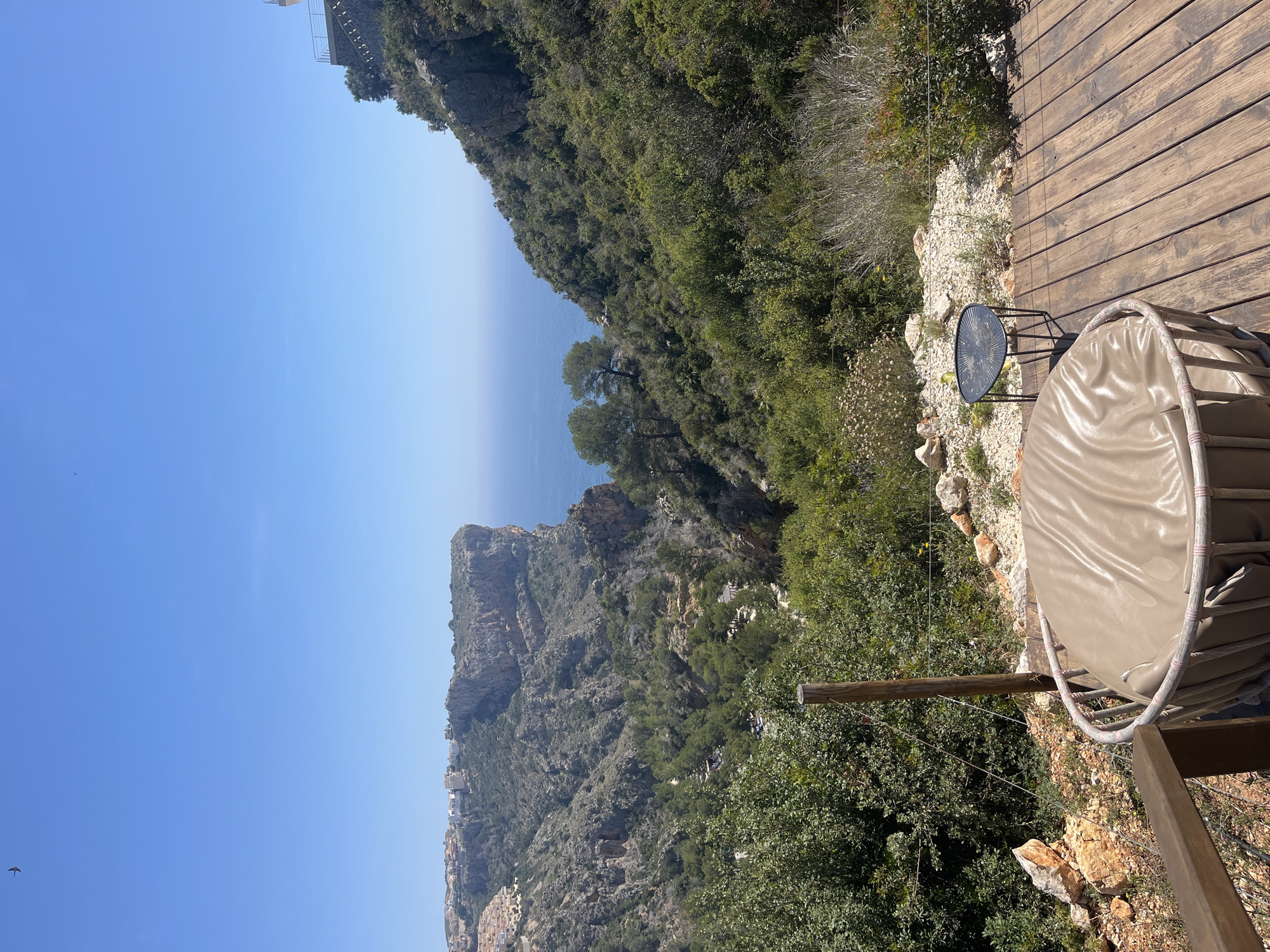 The image shows a view from a wooden deck or patio overlooking the Urbanització El Cim Del Sol area of Spain. In the foreground, there's a round covered seating area and a small round table on a stone-covered portion of the deck. Beyond the deck is a cliff covered in dense green vegetation. In the distance is the ocean, and further away is a rocky, mountainous landscape with buildings scattered along the hillside. The sky is blue, and the lighting suggests it's a sunny day.