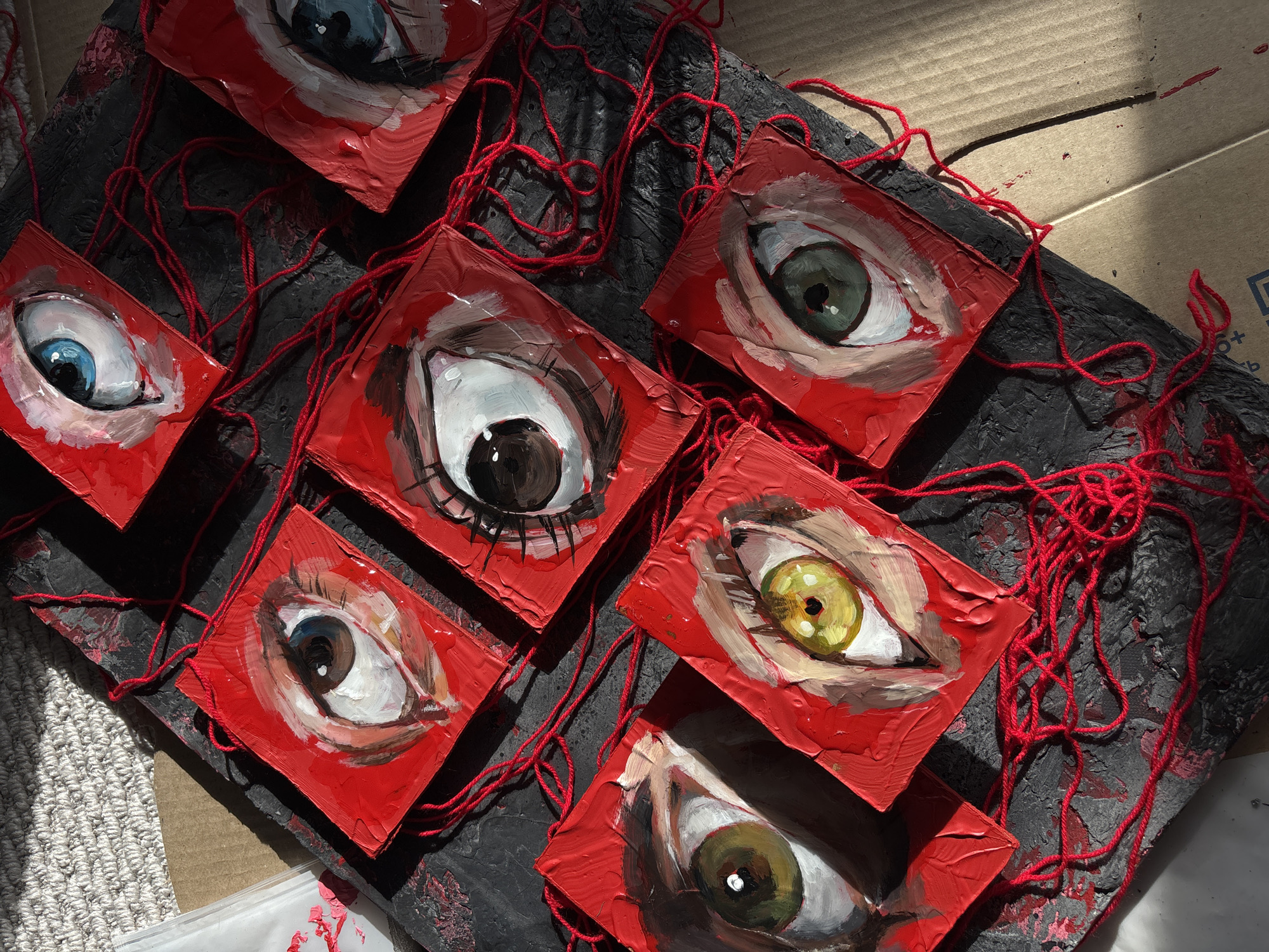 This close-up, high-angle image captures an artistic arrangement featuring multiple painted human eyes, likely set in an indoor space in Krasnoyarsk, Russia.
The primary subjects are eight small, square-shaped art pieces, each depicting a single human eye. These eyes vary in iris color, including distinct shades of blue, green, brown, and black. They are painted with thick, textured brushstrokes on a vibrant red background, giving the paint a glossy appearance in the sunlight. Each eye piece is connected to others by a tangled network of bright red yarn or string, which sprawls across the entire composition.
These eye artworks are displayed on a dark, heavily textured surface, which appears to be a painted board, possibly wood, with an uneven, rough finish. This dark background provides a stark contrast to the bright red of the eye pieces and string. The entire display rests on a lighter brown cardboard surface, visible along the top right and bottom edges, suggesting a workspace or impromptu setup. A light-colored, woven textile or carpet is partially visible in the bottom left corner.
The scene is illuminated by strong, natural sunlight streaming in from the upper right, casting sharp shadows and highlighting the textures of the paint and the dark board. The bright lighting creates a dramatic, high-contrast effect. On the visible cardboard surface, faint blue text and symbols, including a 'B' and a '0+' symbol, are discernible, though not fully legible. The image captures a static artistic composition rather than an activity. The combination of intense eyes and red string against a dark background evokes a somewhat watchful or eerie atmosphere.