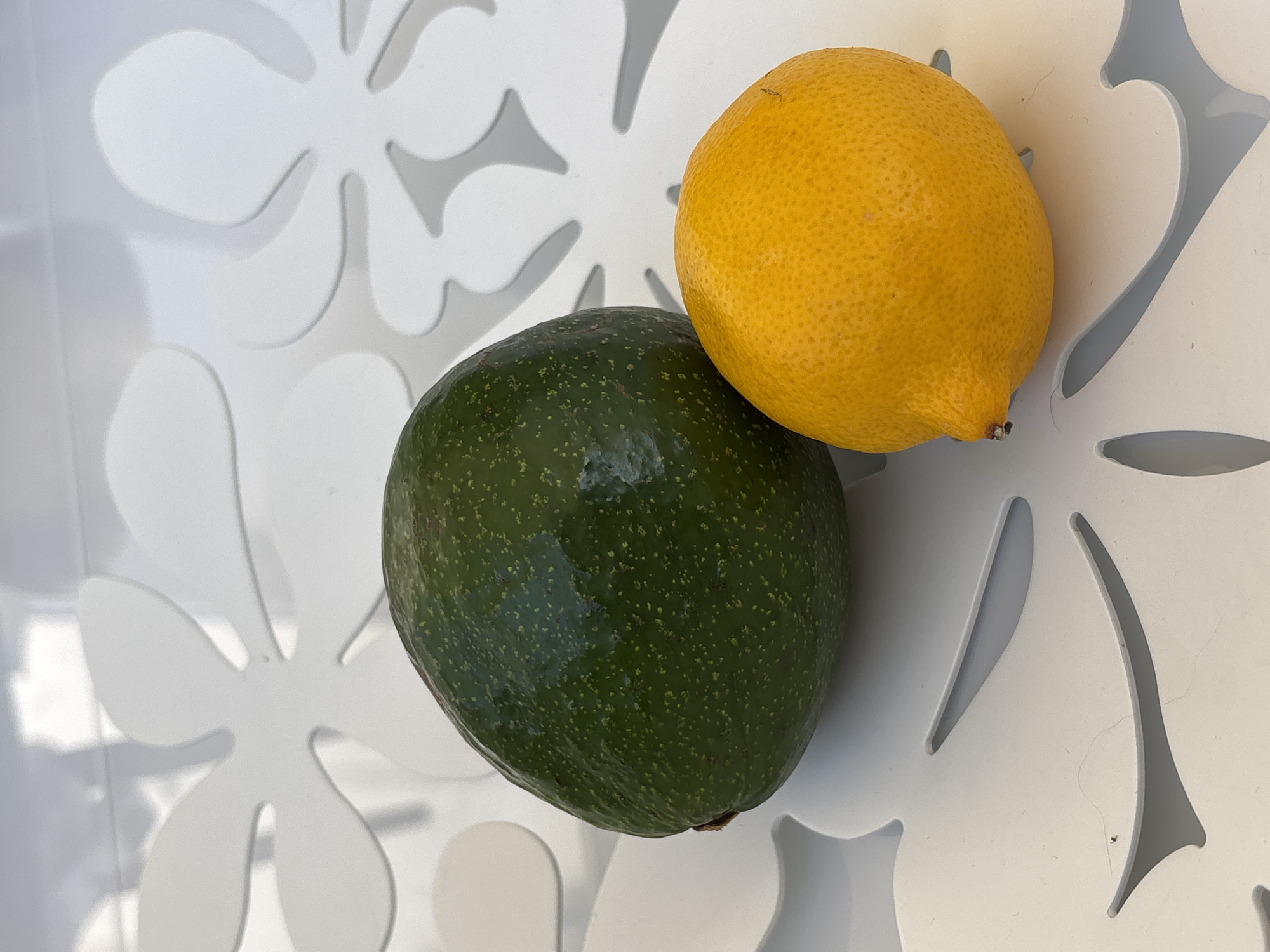 An overhead shot shows a green avocado and a yellow lemon placed on a white surface with a flower-like cutout pattern. The avocado is larger and to the left of the lemon. The lemon is to the right and slightly in front of the avocado. The white surface appears to be a table with a floral pattern. The image is brightly lit, casting shadows on the surface. There are no people or animals in the image. The location is not specified in the image.