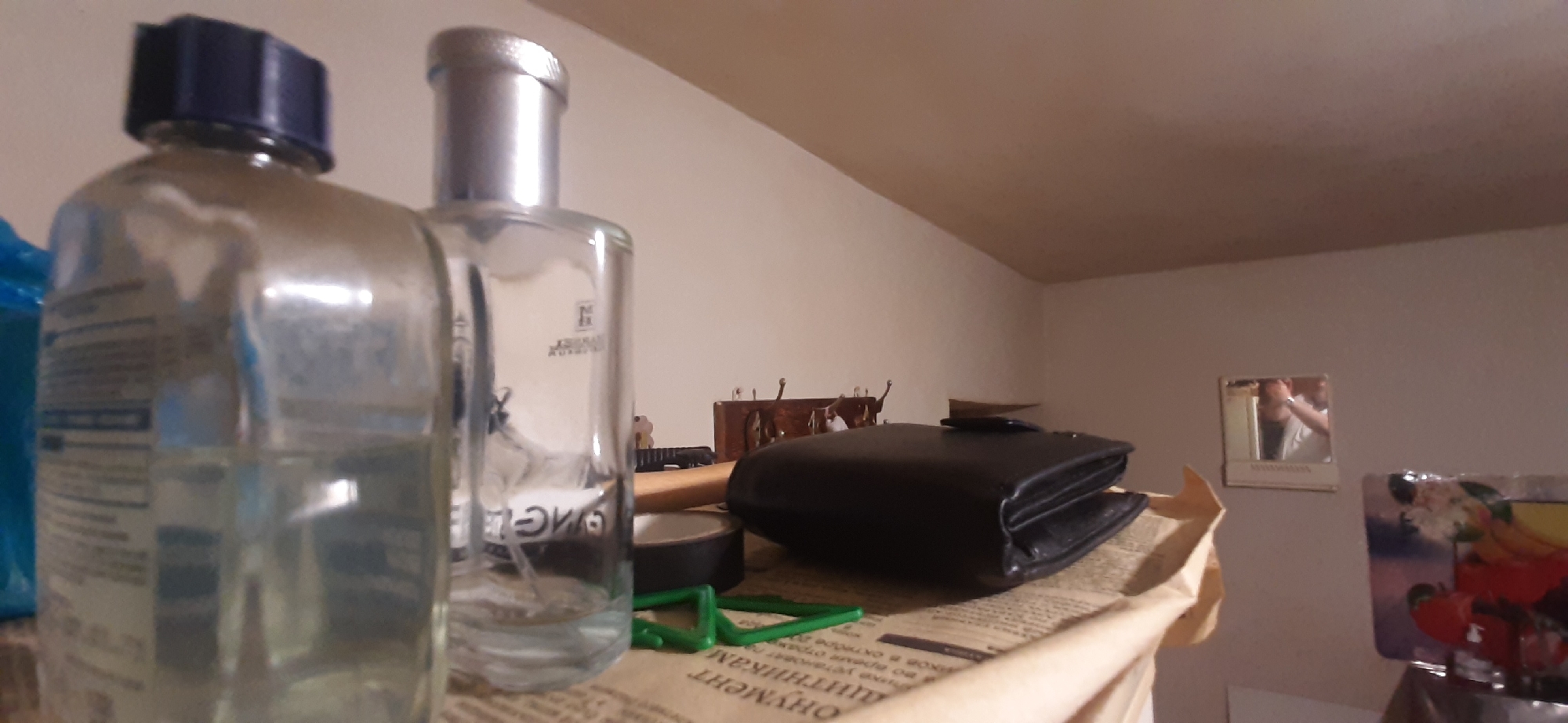 This image depicts a shelf with several items in what appears to be a residential setting. On the left, there is a clear plastic bottle with a blue cap, containing a light-colored liquid. To its right, a taller, clear glass bottle with a silver cap is visible, also containing a clear liquid and bearing some text on its side. In the foreground, a black leather wallet is placed on top of some papers, which seem to be a newspaper. A roll of black tape and a small green plastic object are also present on the paper. In the background, a wooden coat rack with hooks can be seen, and further to the right, a small square mirror hangs on the wall, reflecting a person. Another framed object with colorful imagery is partially visible on the far right. The overall lighting suggests an indoor environment, possibly a room or a closet. The location is identified as Yessentukskaya, Russia.