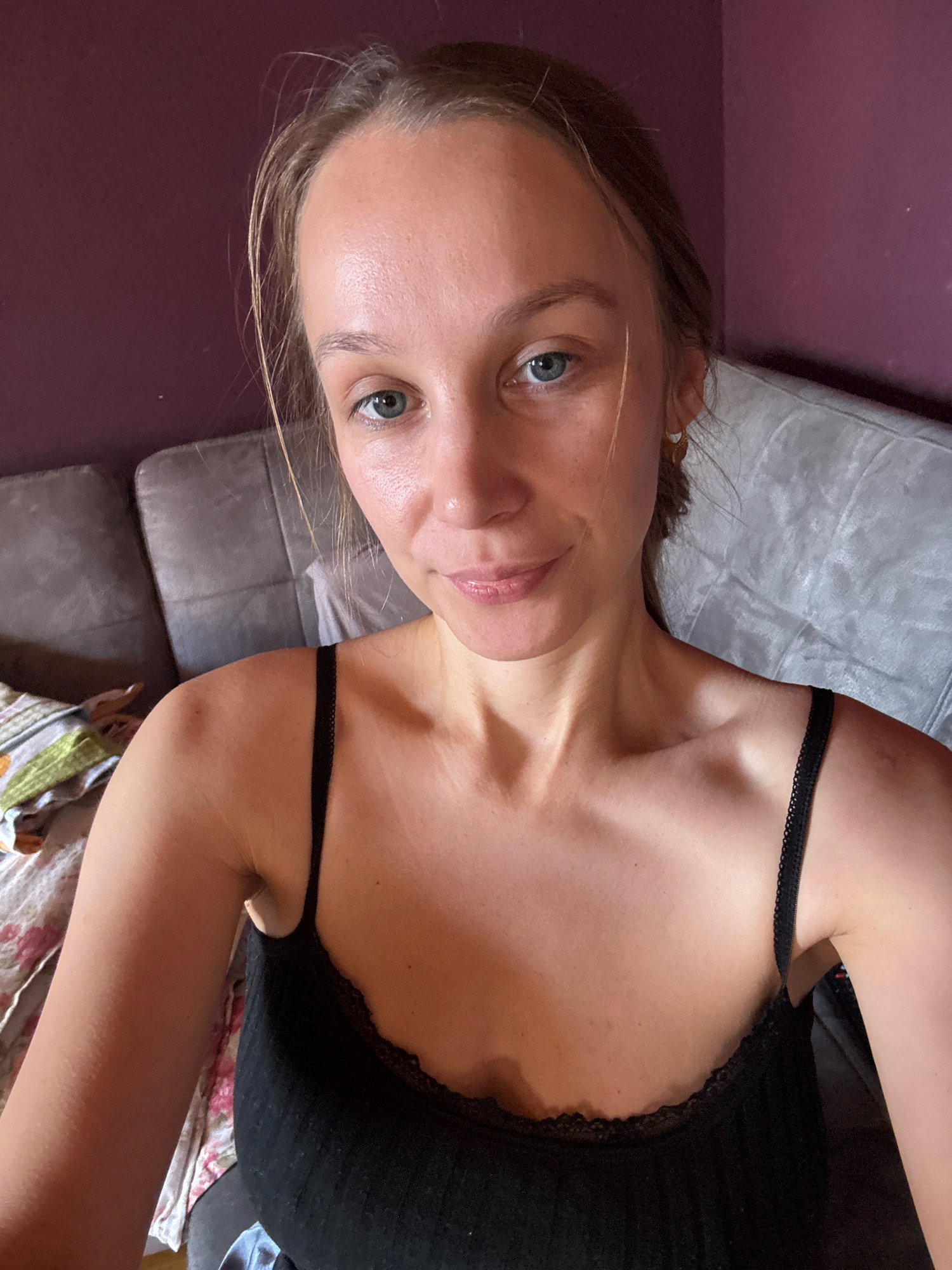 A young woman with blonde hair and blue eyes is pictured from the chest up, looking directly at the camera. She is wearing a black tank top with lace trim. Her skin appears smooth with a slight sheen. She is positioned in front of a grey sofa and a purple wall. The background is slightly out of focus, with some patterned fabric visible on the left side of the frame. The lighting suggests an indoor setting, likely a living room. There is no information or visual evidence to suggest the location is Überherrn, Germany.