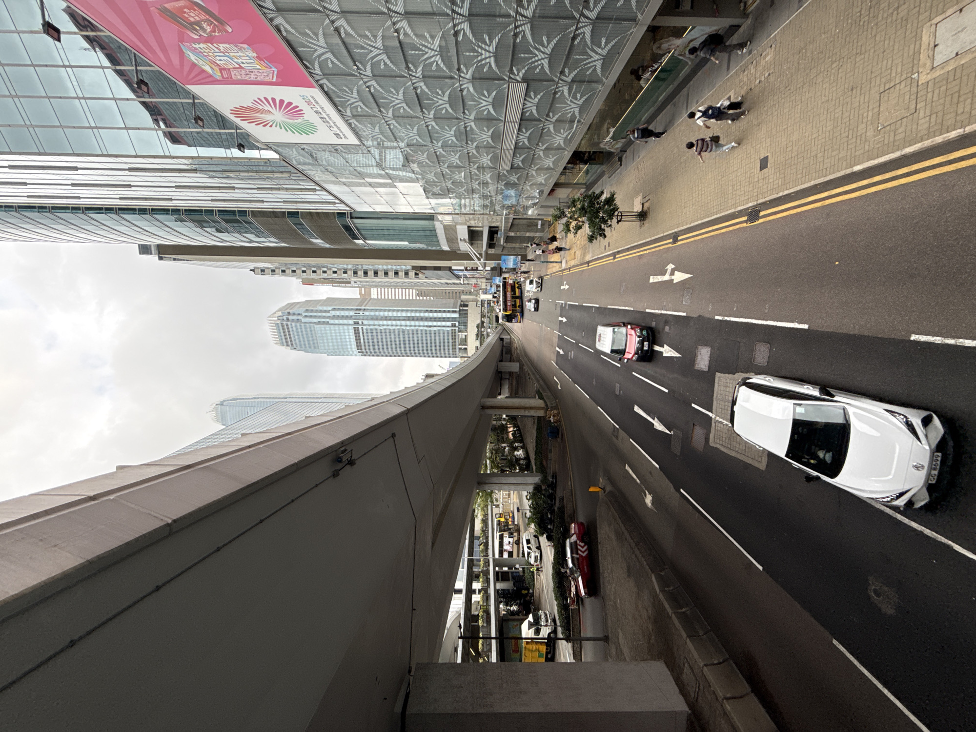 This is a street scene in Hong Kong, China, viewed from an elevated position, possibly a walkway or bridge. The street is divided by a median and has multiple lanes of traffic. Two cars are visible, one white and one red/black. Pedestrians are walking on the sidewalk on the right side of the frame. Overhead, there are tall buildings with glass facades. One building has decorative patterned glass and a pink and white banner. A concrete structure, possibly a pedestrian bridge, dominates the lower left corner of the frame. The sky is overcast.