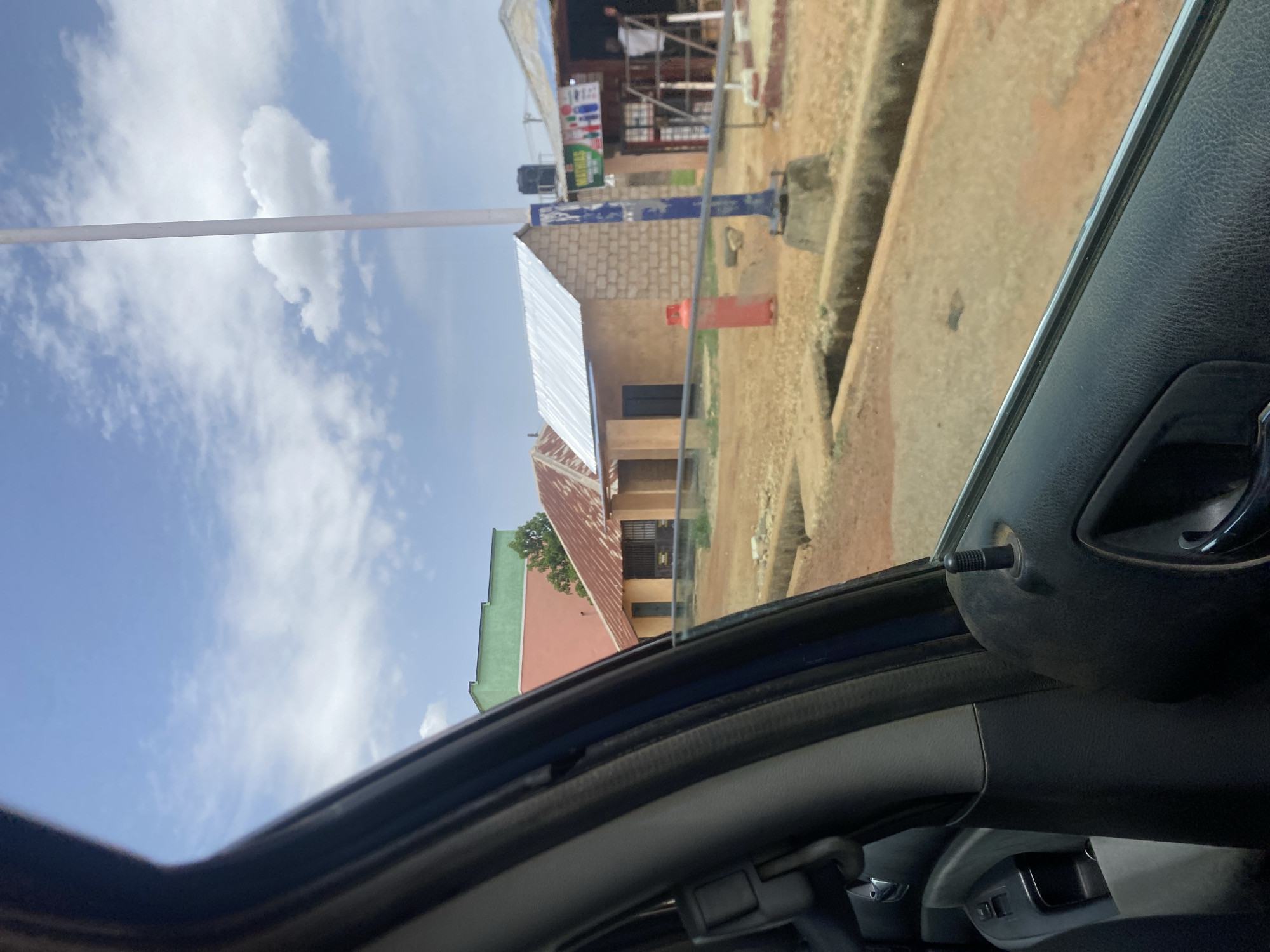 The image depicts a view from inside a vehicle, looking out onto a street in Jos, Nigeria. The camera is positioned at an angle, capturing parts of the car interior (door panel, window frame, and interior roof). Outside the car, there are buildings of various construction types, including brick and sheet metal roofs. Some buildings appear to be shops or businesses. A utility pole is visible in the middle ground of the image. The sky is blue with scattered white clouds. The overall scene is brightly lit, indicating daytime.