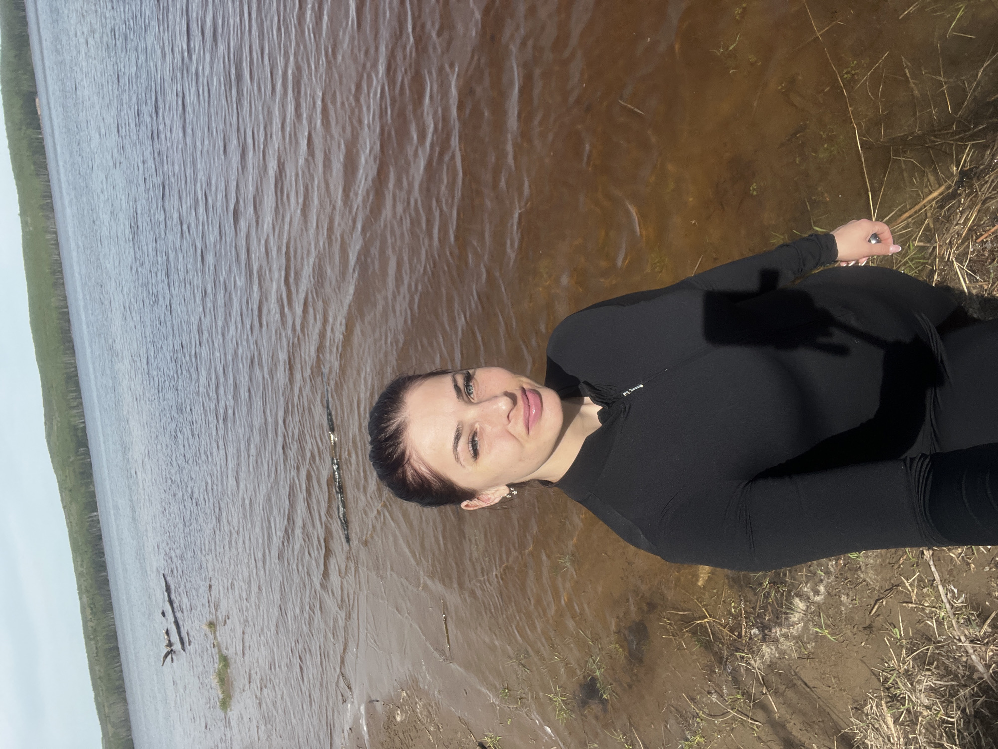 A woman stands on the shore of a body of water. She is wearing a black, long-sleeved garment. The water appears murky and brown, with some vegetation visible at the shoreline. A line of trees is on the opposite shore. The location is Chaikovsky, Russia.