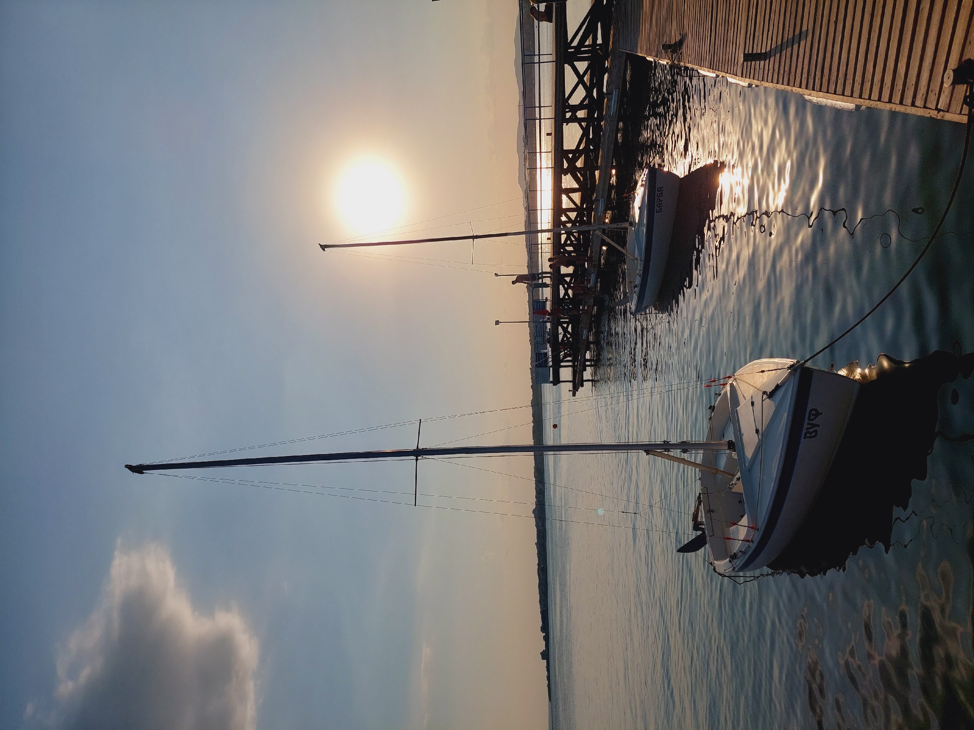 The image depicts a serene sunset scene at a marina in Nesebar, Bulgaria. The primary subjects are two sailboats docked along a wooden pier. The foreground features a sailboat with its mast prominently displayed, extending towards the clear sky. In the background, another sailboat is moored near the pier, partially obscured by the structure. The pier itself is constructed of weathered wooden planks, reflecting the warm glow of the setting sun.
The activity taking place appears to be a calm evening at the marina, with no people actively engaged in any specific task, suggesting a moment of quiet repose. The time of day is clearly sunset, indicated by the low position of the sun and the warm, golden light casting long shadows and creating shimmering reflections on the water. The weather seems pleasant, with a clear sky except for a few wispy clouds, contributing to the tranquil atmosphere.
Notable details include the way the sunlight dances on the water's surface, creating a rippling effect. The wooden pier shows signs of age and exposure to the elements, adding a rustic charm to the scene. The background reveals a distant coastline with faint outlines of hills or mountains. The emotions evoked by the image are calmness, peace, and the beauty of nature.
Visible text on the sailboats includes markings on the stern of the background sailboat which appear to be Cyrillic letters, possibly the name or registration of the boat. The overall impression is one of a peaceful evening by the sea, a common sight in coastal towns like Nesebar.
