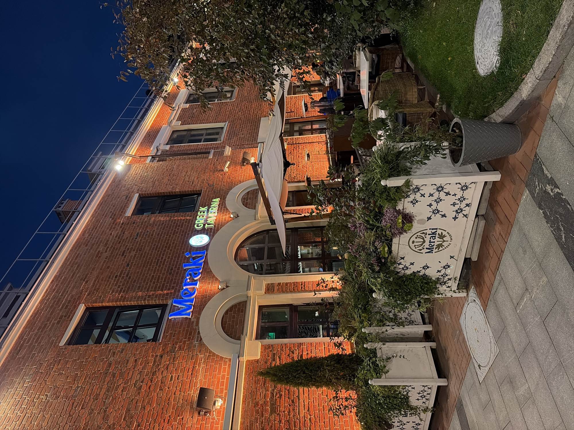 The image shows the exterior of a brick building at night, illuminated by warm exterior lighting. A prominent sign above an arched entrance reads "Meraki GREEK TAVERNA" in blue neon letters. Planters filled with lush greenery and flowers line the sidewalk in front of the building. Wicker chairs and white-topped tables are set up on a patio area, some with white umbrellas providing shade. A gas heater stands on the patio, suggesting cooler weather. A tree with dark green foliage is visible to the right. The ground is paved with cobblestones and modern grey tiles, with a manhole cover visible on the tiled section. The overall atmosphere is inviting and suggests an outdoor dining area for a restaurant. The image is taken in Moscow, Russia, specifically at "Meraki Greek Tavern" located at 20 Bolshaya Nikitskaya Street, Moscow, 125009, Russia.