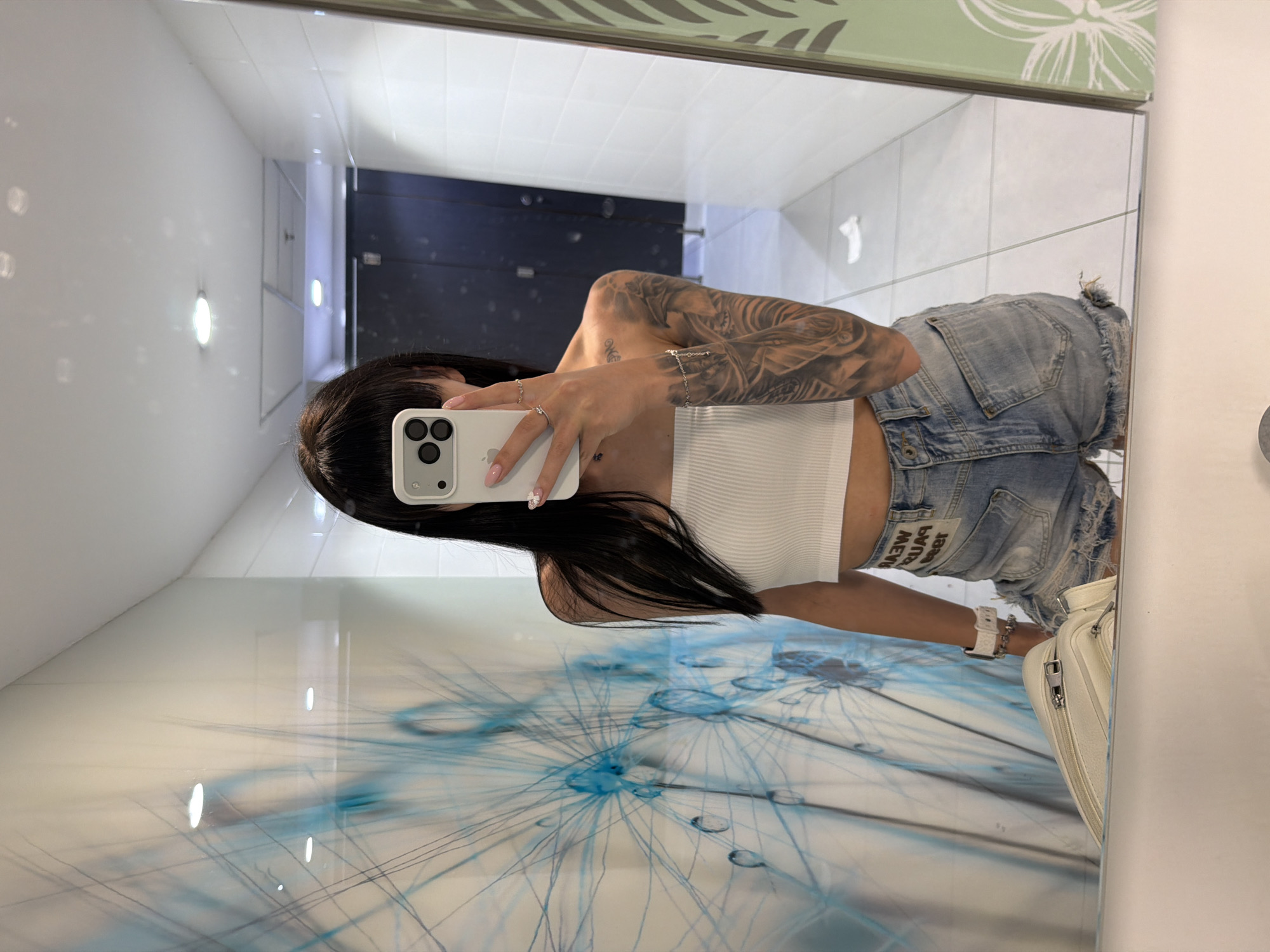 The image depicts a young adult female taking a mirror selfie in what appears to be a modern public restroom, identified as being within Mall Plovdiv, Plovdiv, Bulgaria.
The subject has long, dark hair with bangs and is wearing a white ribbed bandeau crop top and light blue distressed denim shorts. Her left arm is extensively tattooed with a full sleeve of intricate designs. Her right wrist displays a white smartwatch and two bracelets, one metallic beaded and one silver chain. She wears a ring on her right ring finger and another on her right middle finger, and her fingernails are painted light pink. She holds a light grey iPhone with three camera lenses in her right hand, obscuring most of her face as she photographs her reflection.
The environment, visible in the mirror's reflection, features white tiled walls. The wall to the viewer's left is adorned with a large, distinctive blue and white mural depicting a dandelion-like structure with water droplets. The wall to the viewer's right is plain white tiled. The ceiling is white with multiple recessed circular light fixtures. Reflected in the background are dark doors, likely indicating restroom stalls. The floor consists of light-colored tiles, and a crumpled piece of white paper or tissue is visible on the floor near the right wall. A portion of the mirror's frame on the far right edge is light green with white floral or leaf patterns. A white structured handbag or purse rests on a counter or shelf surface in the lower foreground.