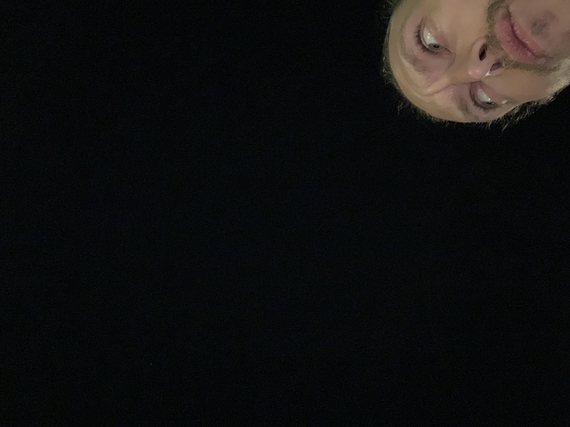 A close-up shot of a person's face in the upper right corner against a dark, seemingly black background. The person has light skin, visible eyes, nose, and lips, and appears to have a short beard or stubble. The facial expression is ambiguous. The background is undifferentiated and appears to be entirely dark. There is no indication of a specific location in Tel-Aviv, Israel, or any other city. The scene lacks context.