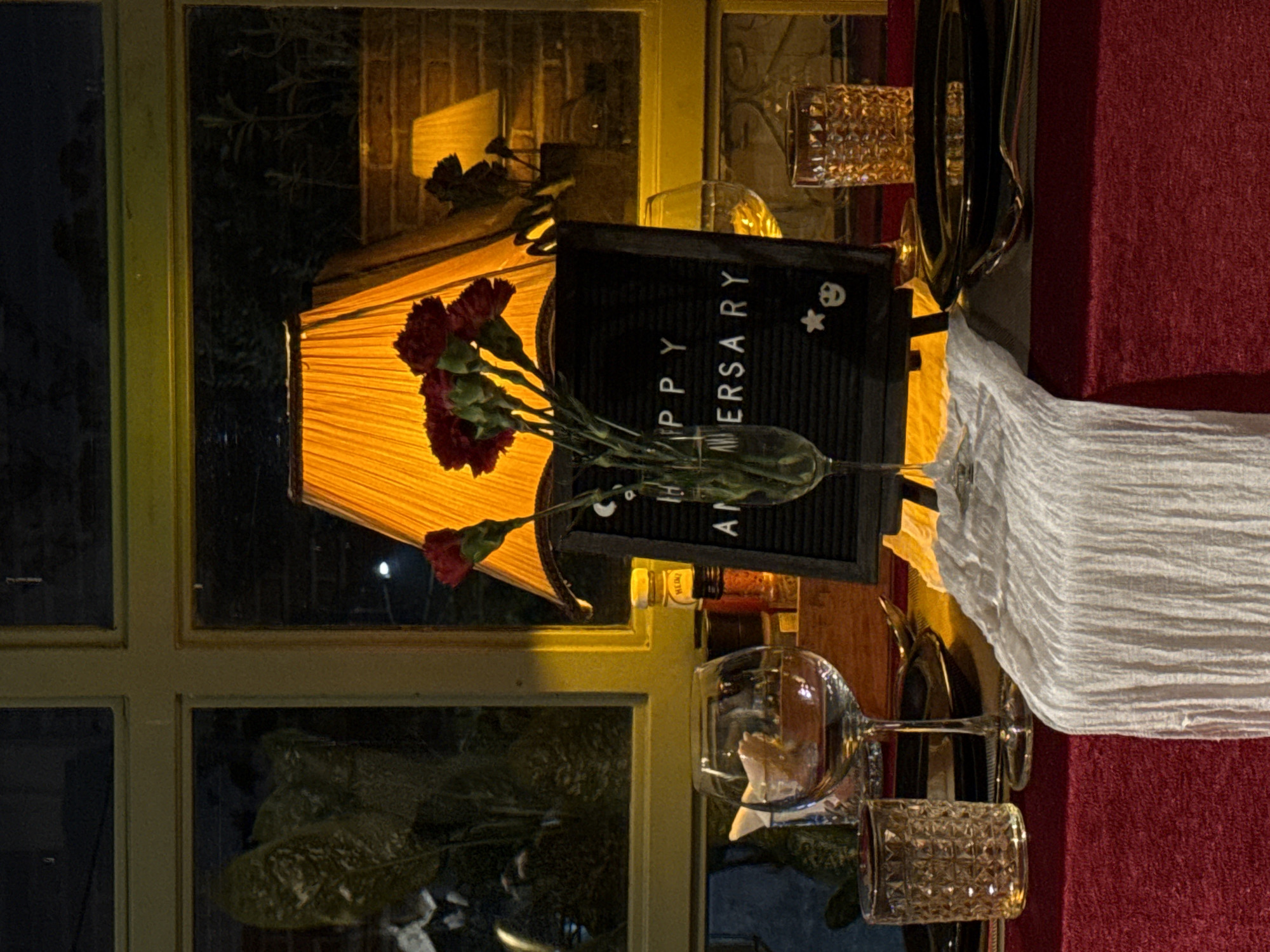 The image shows a festive table setting, likely indoors near a window. A lamp with a yellow shade illuminates a "Happy Anniversary" sign, placed near a vase of red carnations. Two wine glasses, along with what appears to be a decorative tumbler, are also on the table. A white fabric drape spills onto a red surface. The setting evokes a romantic or celebratory atmosphere, perhaps a private event. The location is not explicitly clear from the image but based on the information given, it takes place in the city of Lâm Viên - Đà Lạt, Vietnam.