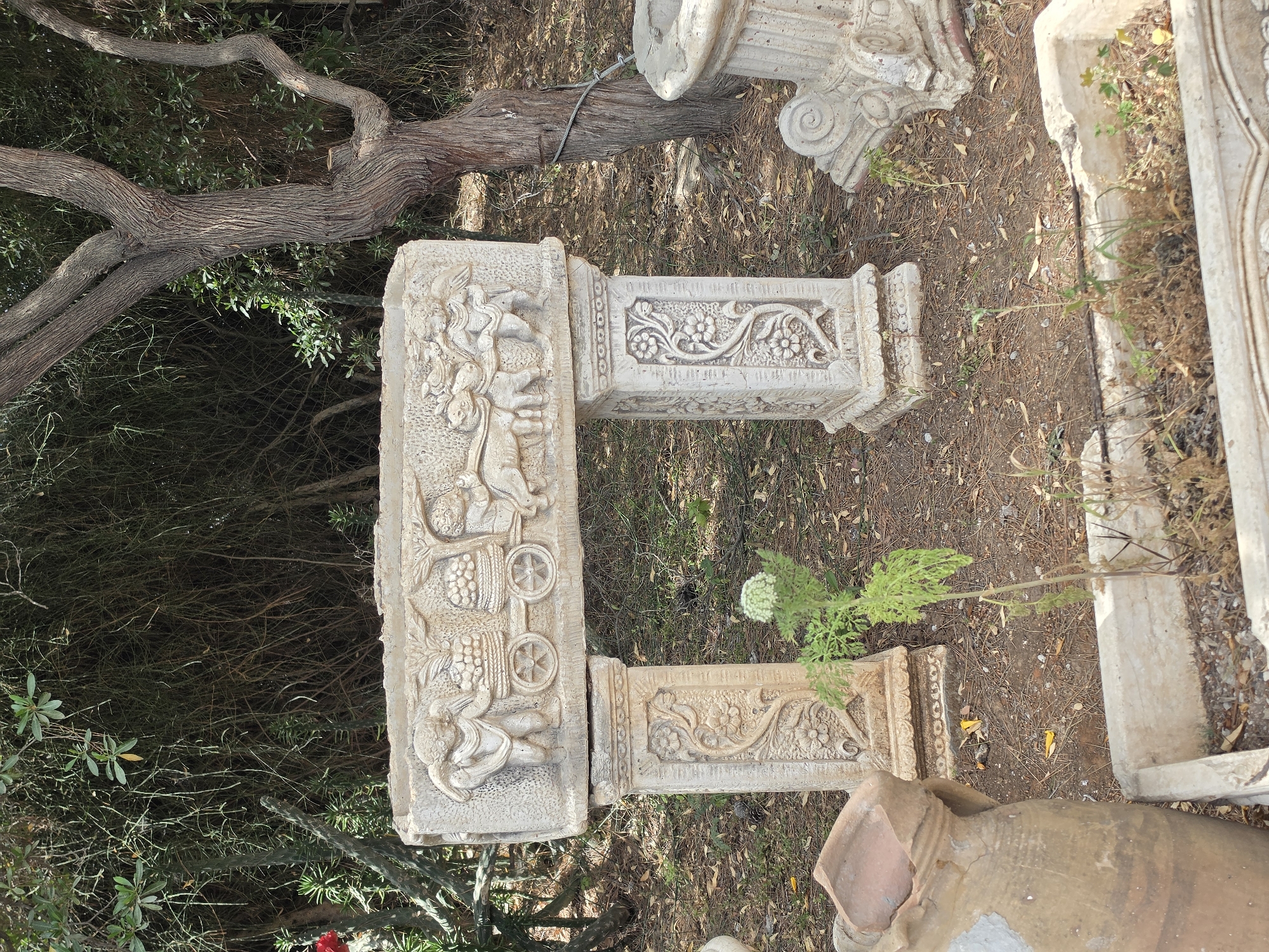 The image shows a collection of weathered, decorative stone objects in an outdoor setting, likely in Mate Yehuda Regional Council, Israel, suggested by the landscape characteristics. The objects consist of sculpted stone blocks with intricate carvings, possibly fragments of architectural structures. The carvings depict figures and floral designs, indicating an origin from a decorative or artistic context. The stones are placed among dry leaves, scattered vegetation, and near a large tree branch, possibly in a garden or an area with neglected artifacts. The presence of additional fragments and a clay vessel suggests the location is a storage area for discarded architectural pieces. There are no people in the scene.
