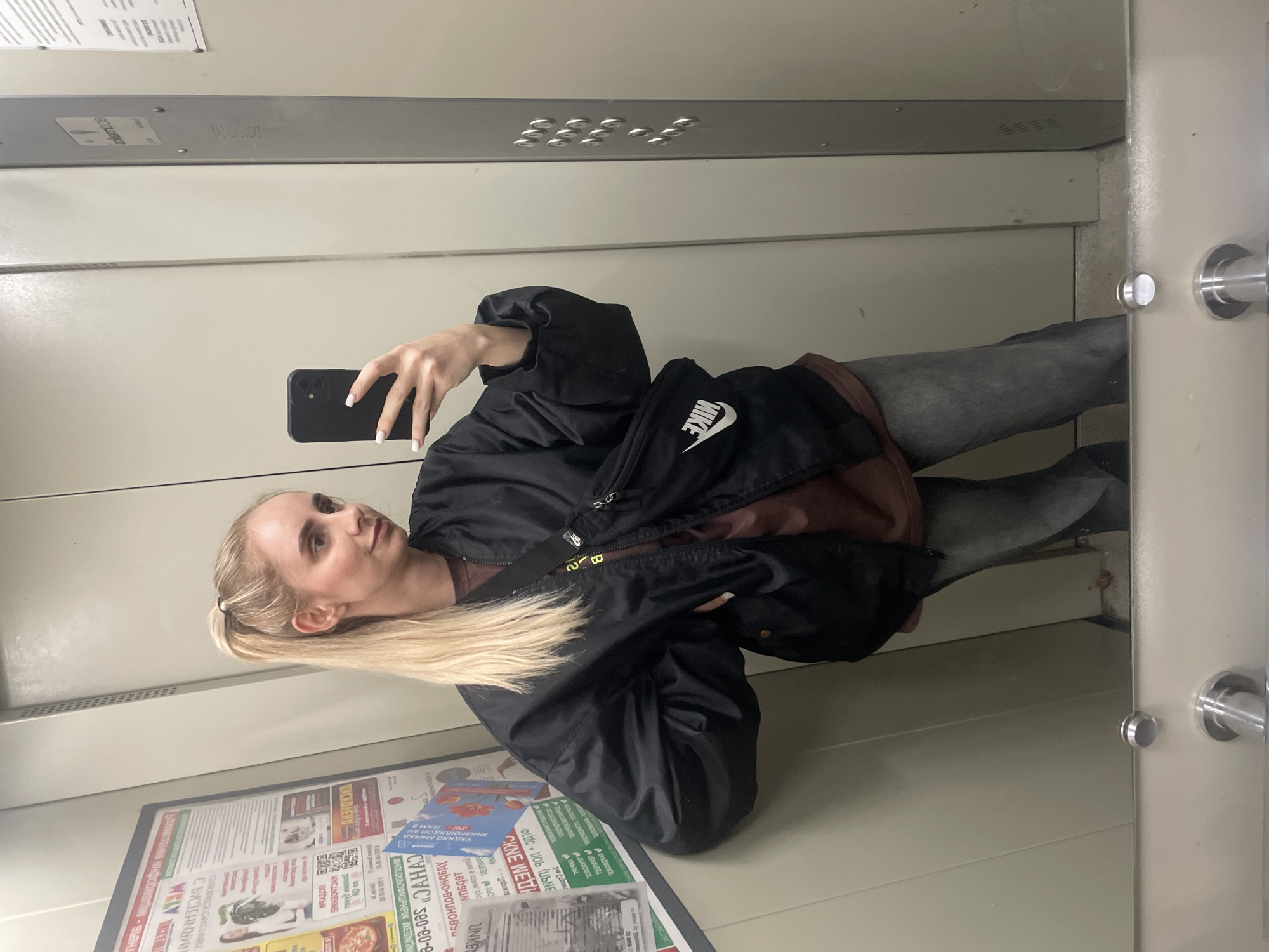 A woman with blonde hair pulled back in a ponytail is taking a selfie in what appears to be an elevator. She is holding a black smartphone. She is wearing gray pants, a brown shirt, and a black Nike jacket. On the walls of the elevator are posters, including one that reads "Vladivostok" in Russian.