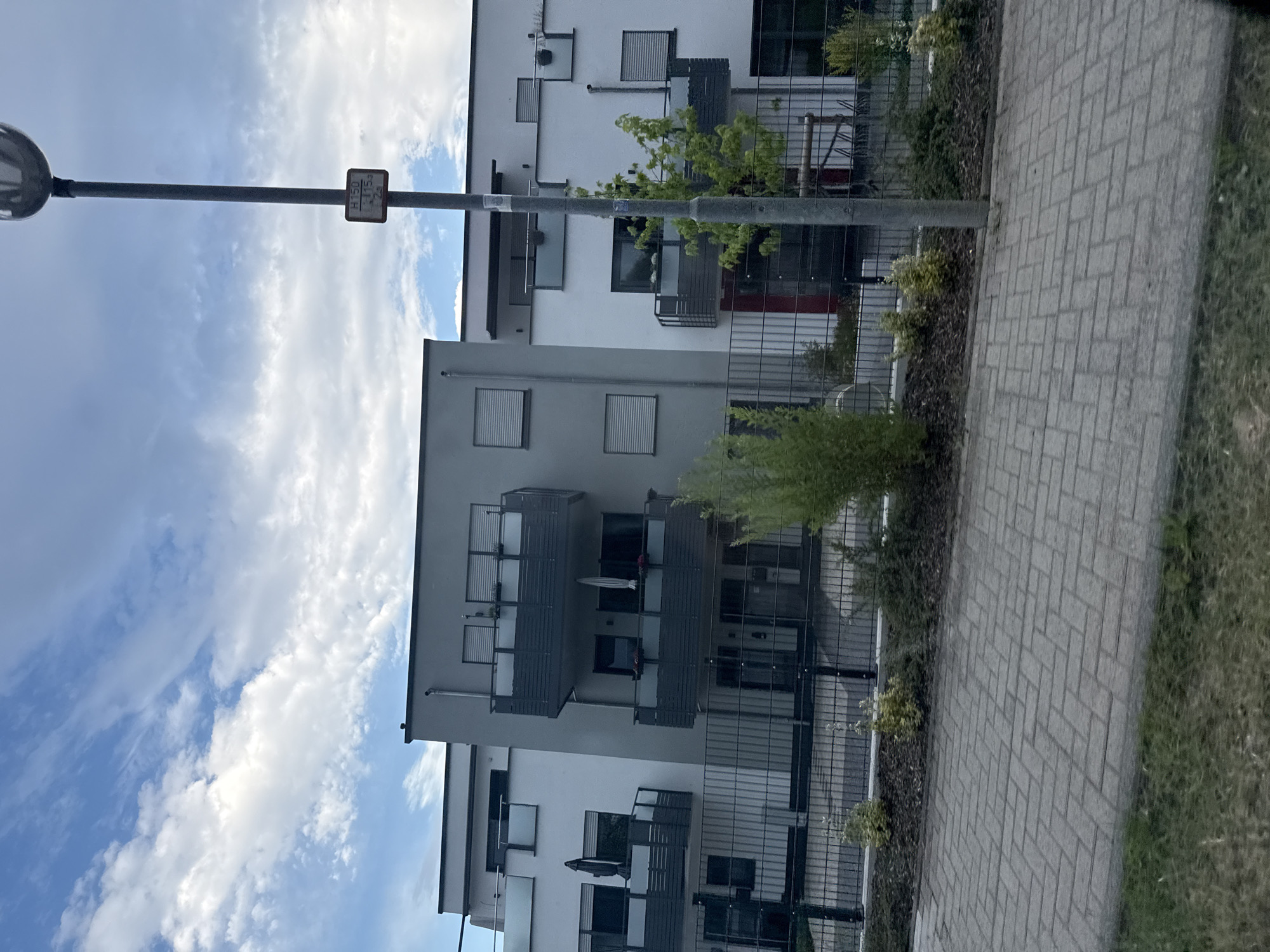 The image shows a modern apartment building in Volpertshausen, Germany, with a cloudy sky above. The building features several balconies with dark railings and some greenery on them. A paved walkway runs in front of the building, separated by a black metal fence and a strip of landscaping with small trees and bushes. A lamppost with a sign is visible to the right of the building. The architecture is contemporary, with clean lines and a mix of white and grey facades. The overall impression is that of a well-maintained residential area.