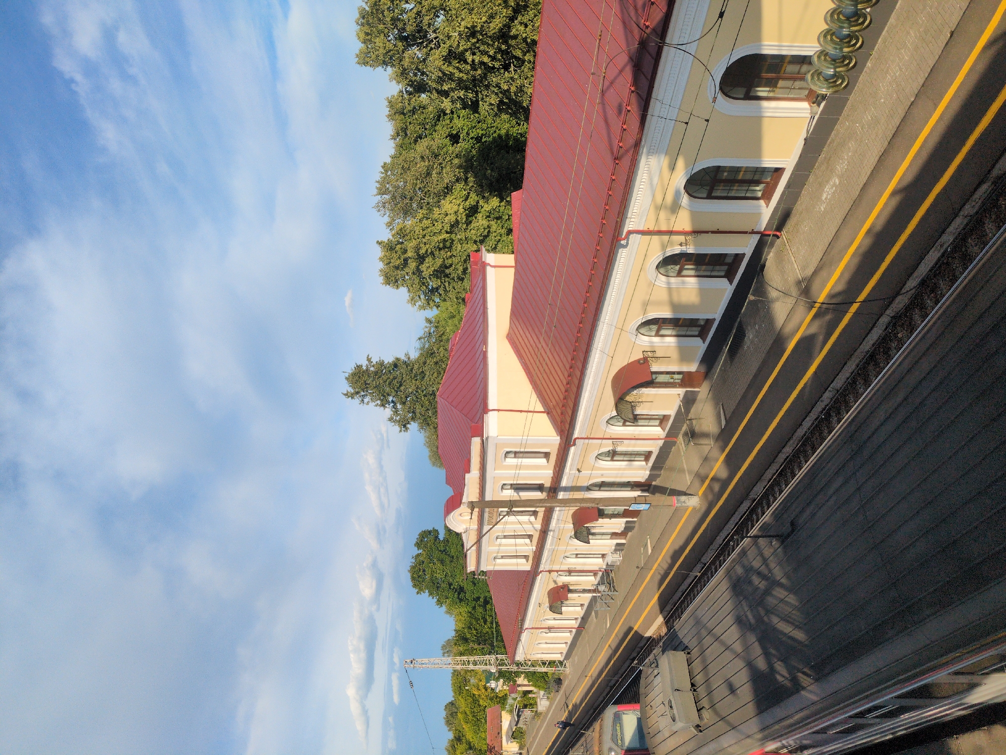 The image shows a train station in Velikovo, Russia. The station building is a long, light yellow structure with a red roof and arched windows. There are awnings over some of the windows and doors. A train is parked at the station on the tracks next to the platform. The platform has yellow lines marking the edge. Trees and a clear sky with some clouds are visible in the background.