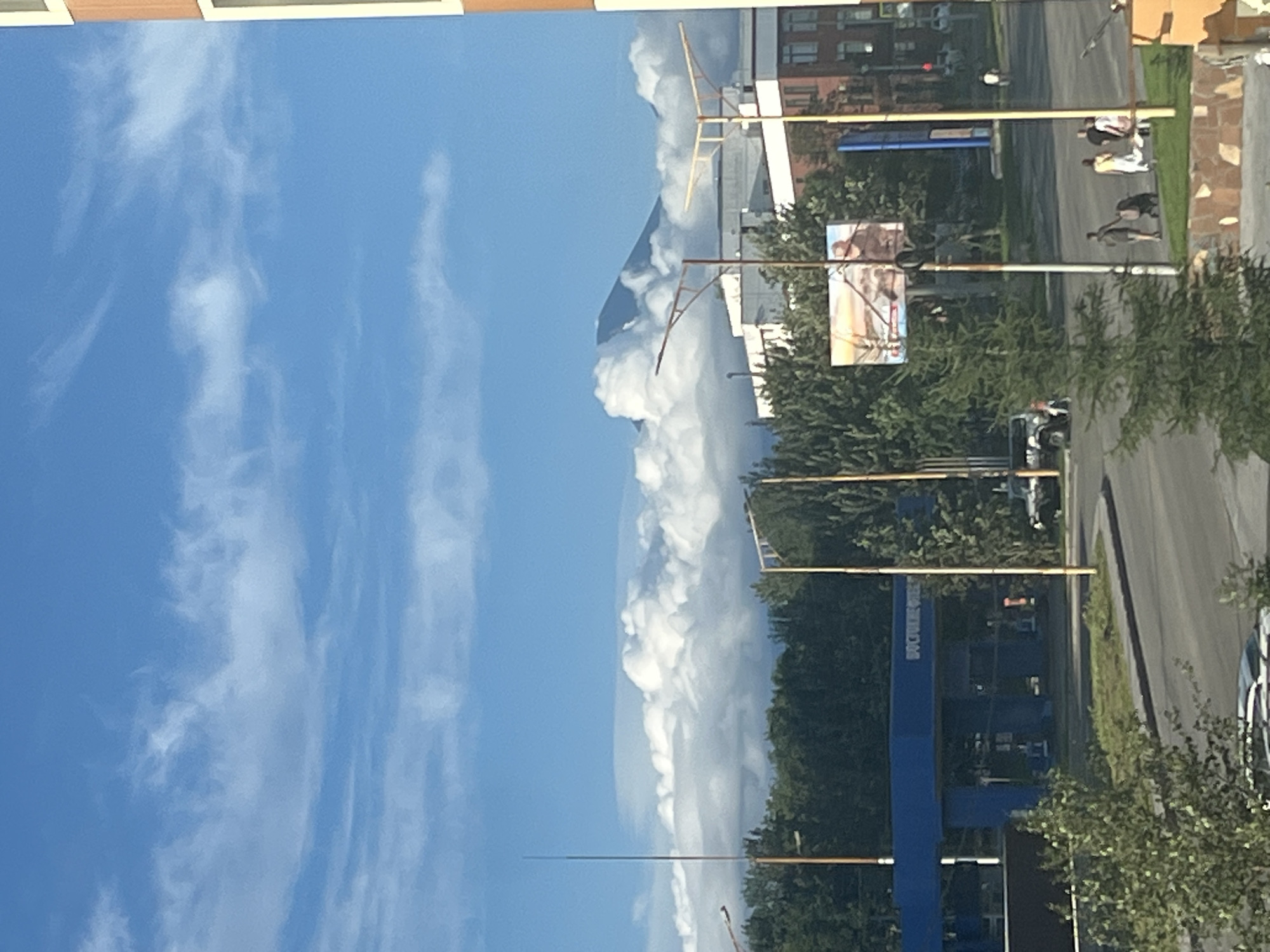 The image captures a street view in Petropavlovsk-Kamchatsky, Russia, with a large, snow-capped mountain peak visible in the distance, partially obscured by clouds. In the foreground, a blue gas station canopy stands prominently, with the inscription "ВОСТОЧНЫЙ" visible on its side. Several streetlights with distinctive yellow poles and angled fixtures are positioned along the road. A billboard advertising some product is situated near the gas station. Trees line the background, and various buildings, including a multi-story brick structure, are visible. People are seen walking along the sidewalk and near the road, with some pushing strollers. Several vehicles, including a white SUV, are also present. The sky is blue with scattered white clouds.