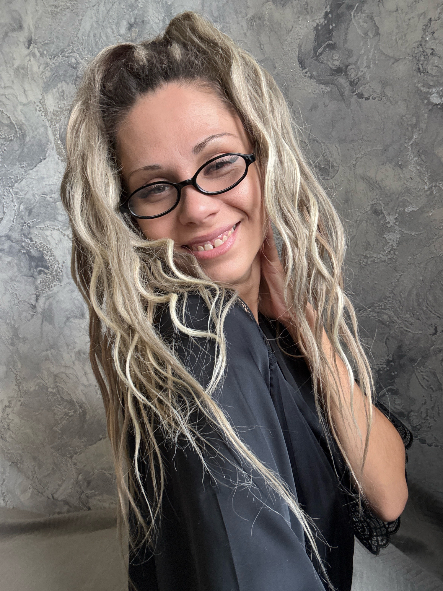 A blonde woman wearing black-rimmed glasses and a black robe is posed against a textured gray wall. Her hair is pulled back into a high ponytail with loose strands framing her face. She is smiling, with her right hand touching her neck. The lighting is soft, creating subtle shadows. The scene appears to be indoors, with no other individuals or prominent objects visible. The location is identified as Rusanovo, Russia.