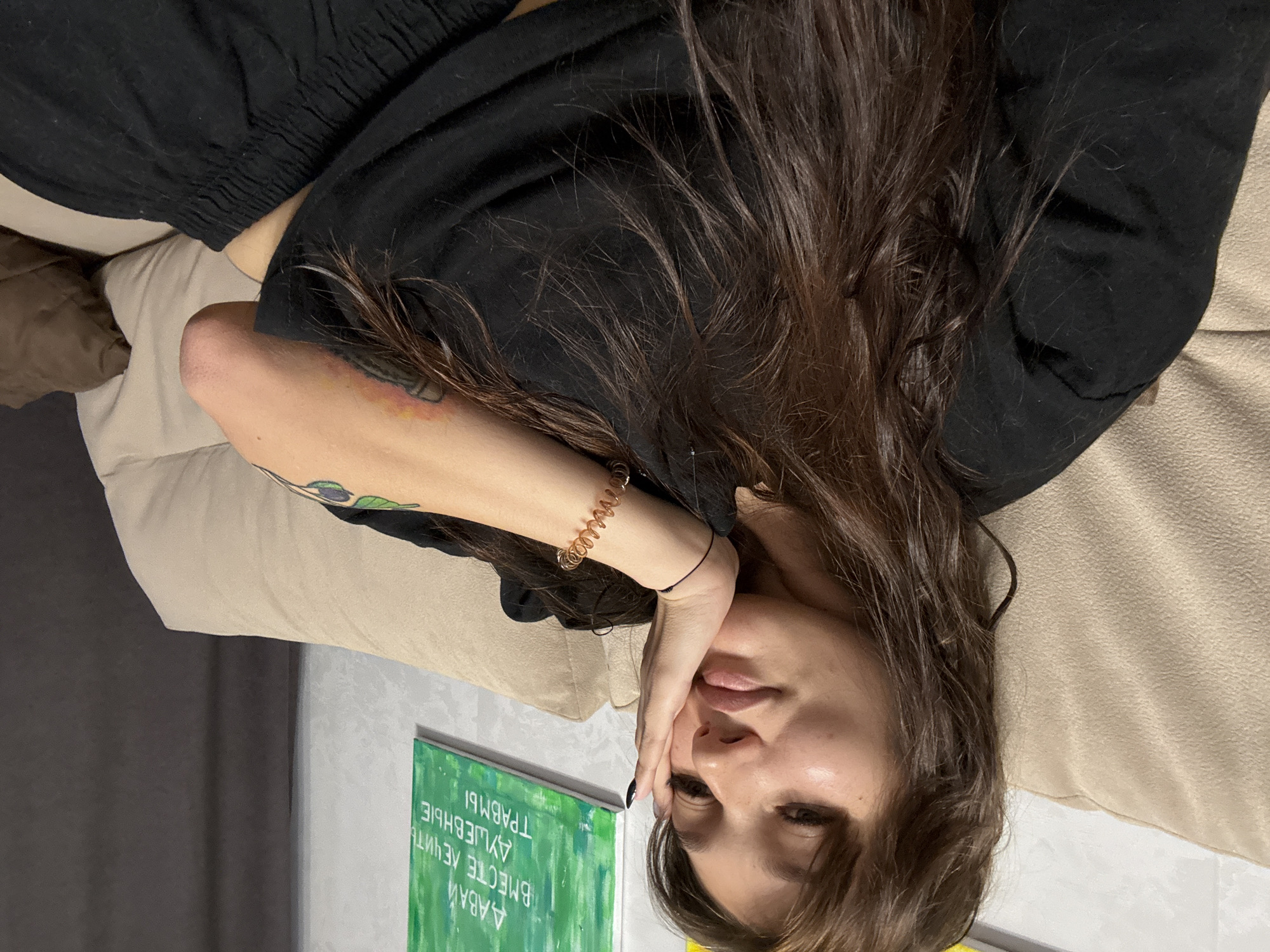 A young woman with long, dark brown hair and bangs is lying on a beige sofa in what appears to be a living room. She is wearing a black t-shirt and black pants. Her right hand is resting on her cheek, and she is looking towards the camera with a soft expression. A copper-colored coiled bracelet is visible on her left wrist. Behind her, on the wall, is a framed green painting with white lettering in Russian. The environment suggests a casual indoor setting, possibly in Novokuznetsk, Russia, given the provided context.