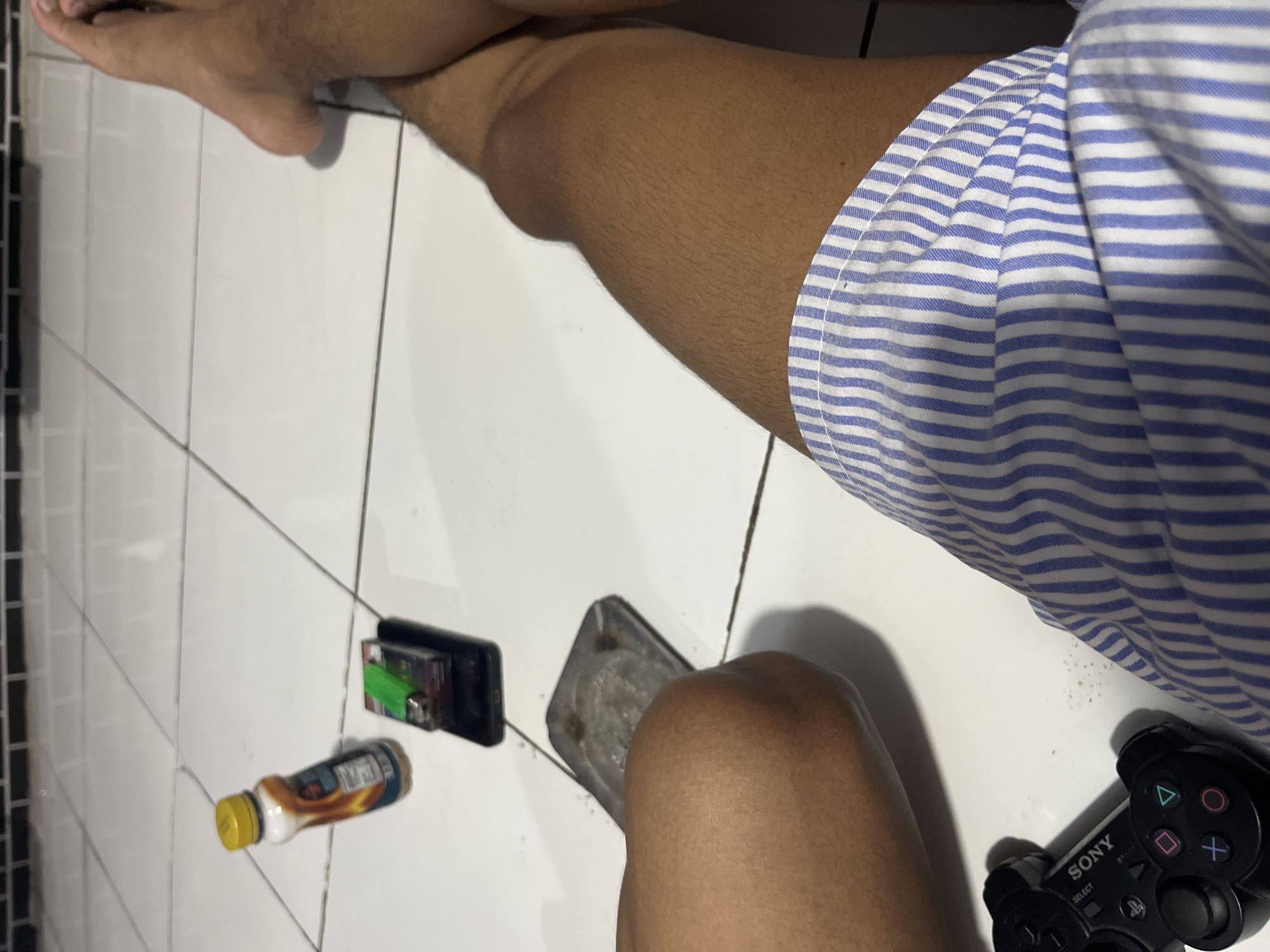 The image depicts a person's legs and torso, seated on a white tiled floor, likely in a residential setting. The individual is wearing blue and white striped shorts. To the left of the frame, a black PlayStation controller is visible, along with a mobile phone topped with a cigarette pack and a lighter. Further to the left, an ashtray containing cigarette butts and a bottle of beverage with an orange and white label are present. The background consists of white tiles with faint grout lines and a dark tiled border at the top. No specific details about the city of Laban, Indonesia are discernible from the image itself.