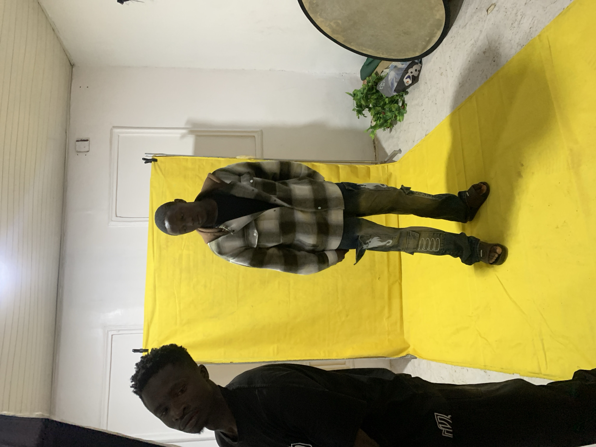 The image depicts an indoor scene, likely a makeshift studio, with a bright yellow backdrop and floor covering. Two young Black men are present. The man on the left, partially visible, is wearing a black t-shirt and black pants with white detailing on the left leg. He is turned away from the camera, with his head tilted. The man on the right is standing centrally in front of the yellow backdrop. He is wearing a plaid shirt over a dark t-shirt, distressed denim jeans with white detailing on the knees and pockets, and dark sandals. He is looking forward, with his hands in his pockets. In the background, to the right, a bundle of green artificial foliage rests on the floor next to a large, circular, reflective object, possibly a studio light reflector. The walls are white, and the ceiling appears to be a white, ribbed material, possibly wood or plastic panels. The overall impression is that of a casual photo shoot or creative session. The setting does not provide any specific visual cues to pinpoint its location within Jos, Nigeria.