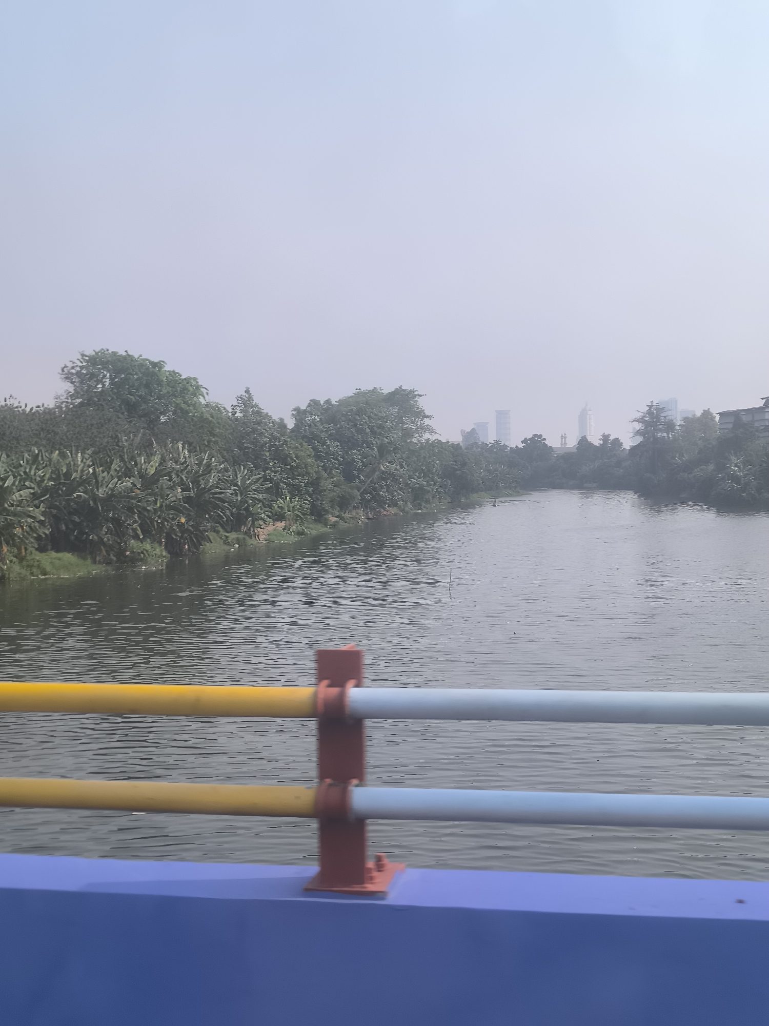 The image shows a wide river flowing through an urban area, with lush green vegetation lining the banks on both sides. In the distance, several modern high-rise buildings rise above the trees, indicating a city skyline. A bridge railing with yellow, blue, and red elements is visible in the foreground, suggesting the photo was taken from a bridge overlooking the river. The sky is hazy and light, creating a diffused light condition. The river surface is rippled, with some debris visible on the water. The overall scene depicts a blend of nature and urban development in Tangerang, Indonesia.