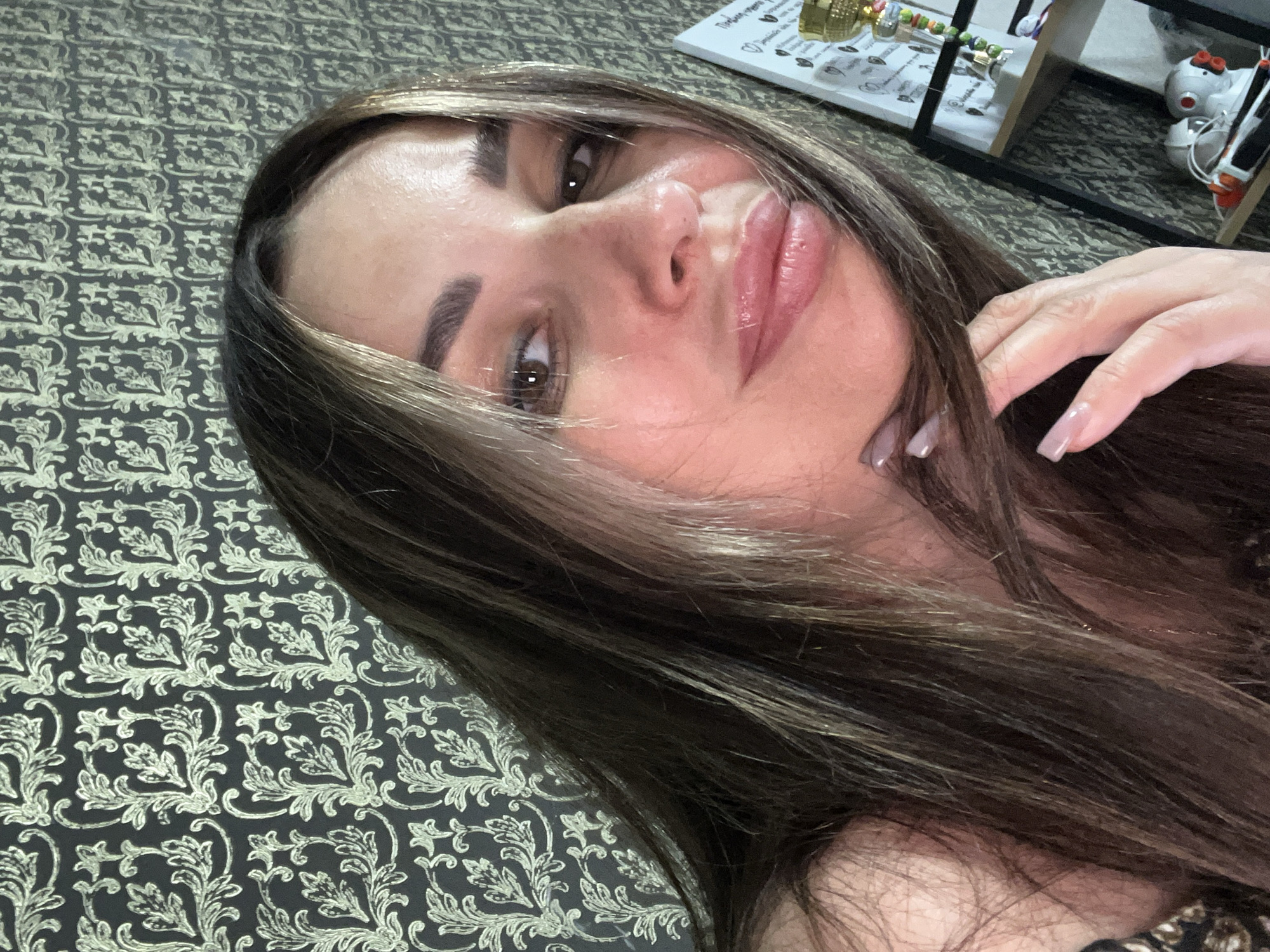 A woman is captured in a close-up, selfie-style shot with her head angled towards the camera, looking over her shoulder. She has long, dark brown hair with lighter streaks, which partially obscures her face. Her eyes are brown, and she is wearing makeup, including eyeliner, eyeshadow, and lipstick. Her lips are slightly parted, and one hand, with manicured nails, is resting near her chin and hair. The background consists of a patterned wallpaper with an ornate, repeating design in a dark color with lighter accents. To the right of the woman, a shelf unit is partially visible, displaying various objects, including what appears to be a trophy or award and some electronic devices. The lighting suggests an indoor setting. The image is cropped tightly around the subject's face and upper body.