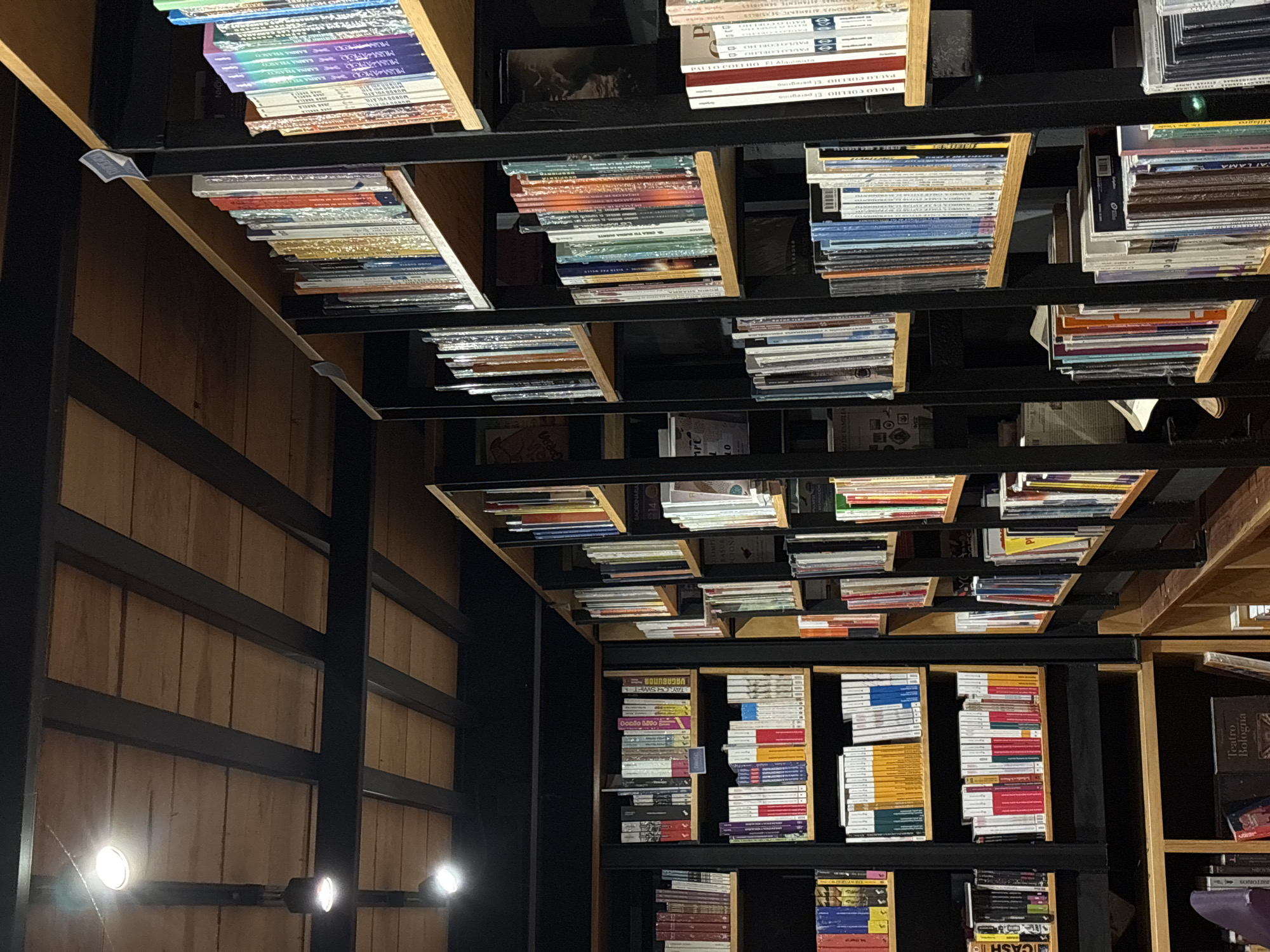 The image shows an unusual bookstore interior, with shelves seemingly built into the ceiling. The books are stacked vertically and horizontally in different sections, with the ceiling shelves giving the impression of a suspended library. The walls are paneled, and track lighting illuminates the scene. Based on the text seen in the image, the bookstore is likely located in Puebla, Mexico. The texts visible include titles such as "Teatro Bologna", "CASH", and "Taylor Swift". The overall environment suggests a unique and perhaps unconventional approach to book storage and display.