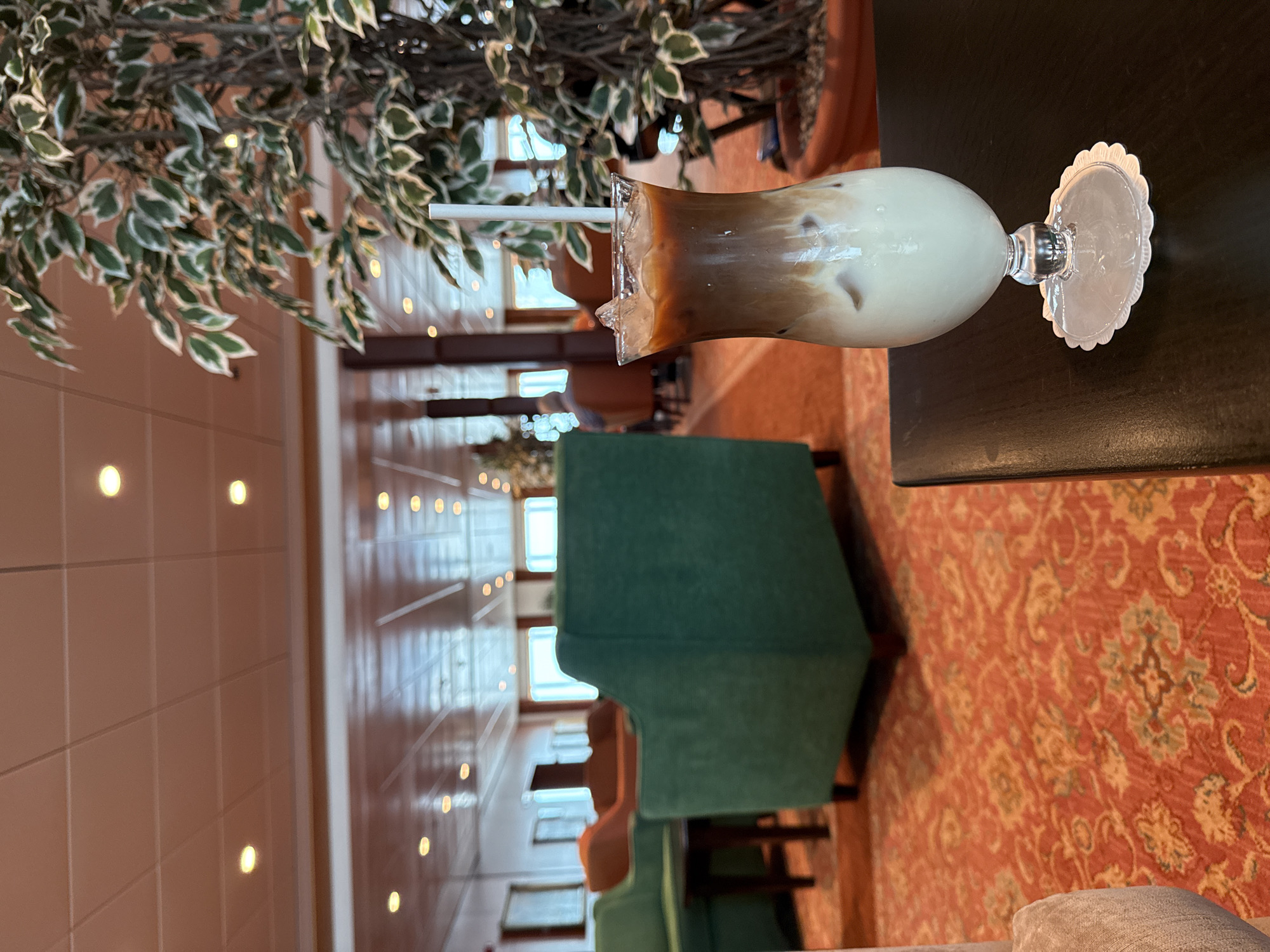 Here is a detailed description of the image:
**Scene:** An interior space resembling a lounge or lobby, potentially of a hotel or upscale establishment, in the city of Unknown, Unknown. The space is decorated with a patterned orange rug and has a row of windows showing bright daylight.
**Objects:**
* A tall glass of iced coffee, partially consumed, sits on a dark brown table. The drink has a straw and is positioned with a decorative coaster.
* A potted plant with variegated green and white leaves is partially visible above the table.
* A green upholstered chair is positioned near the table.
* The background reveals more chairs and tables.
**Environment:**
* The space has an overall warm color scheme due to the rug and interior lighting.
* The window placement suggests the lounge area could be adjacent to a hallway.
**Overall Impression:** The scene presents a quiet, relaxed environment suitable for casual meetings or a quiet break.