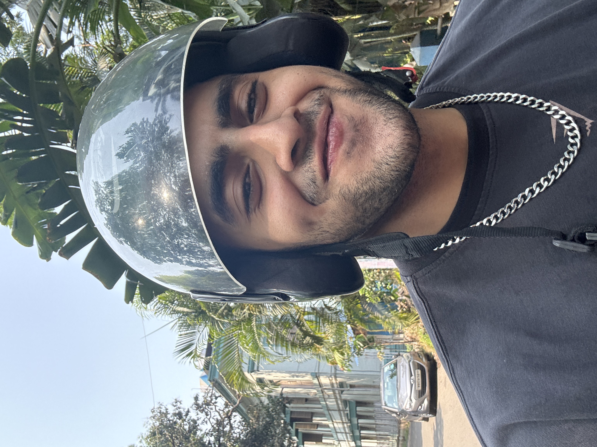 A medium shot captures a man wearing a black t-shirt, a silver chain, and a reflective helmet. The background includes a cityscape, palm trees, buildings, and a car visible on the street. The photograph was taken in Kolkata, India.