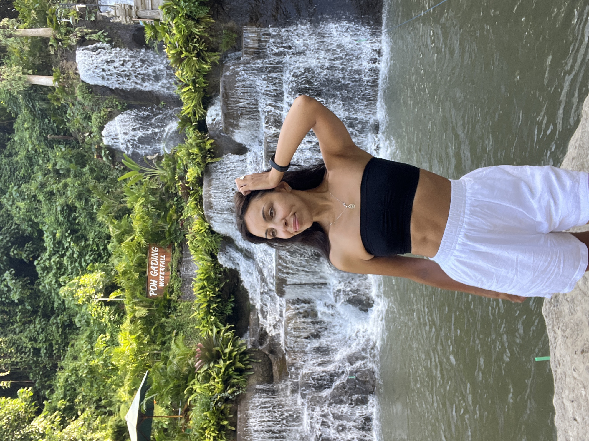 A person, likely female, stands beside the Poh Gading Waterfall, located in Sukawati, Indonesia. She has dark hair and fair skin, wears a black bandeau top, and white shorts. The waterfall cascades down a series of rocky tiers surrounded by lush greenery. Water flows into a pool. A sign with the words "POH GADING WATERFALL" is visible.