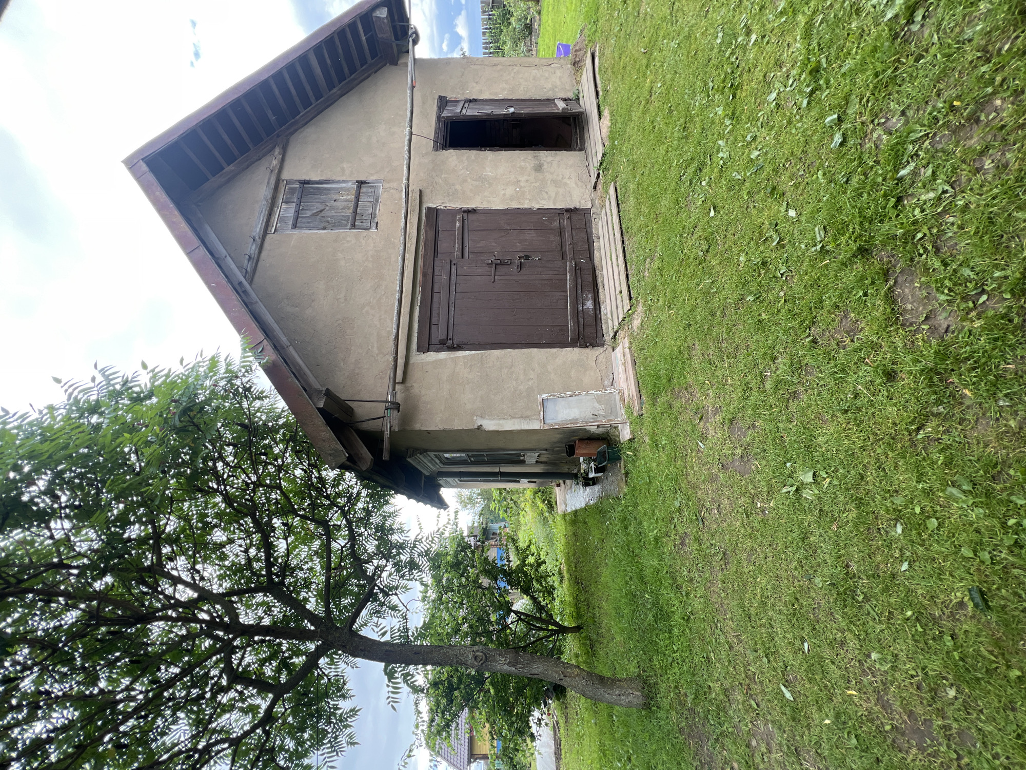 This image depicts a rustic, weathered barn or outbuilding in a rural setting, likely within the Бучацінскі сельскі Савет of Belarus. The structure is characterized by its rough, beige plastered walls and a steeply pitched, rust-colored metal roof. A wooden window with shutters is visible on the upper facade. The main entrance features large, dark brown double doors with visible hardware and a padlock. To the right of these doors is a single, smaller wooden door, partially ajar, revealing darkness within. Both entrances are fronted by wooden planks serving as makeshift walkways over the grassy ground.
The barn is situated on a patch of uneven, green lawn, with scattered leaves and patches of bare earth. To the left of the barn, a mature tree with a thick trunk and leafy branches casts some shade. In the background, other rural dwellings and vegetation are visible, suggesting a residential area. The sky above is overcast with some white clouds. Various small objects, including pots and a discarded window frame, are scattered near the side of the barn. The overall impression is one of an aging, functional outbuilding in a quiet, rural Belarusian landscape.