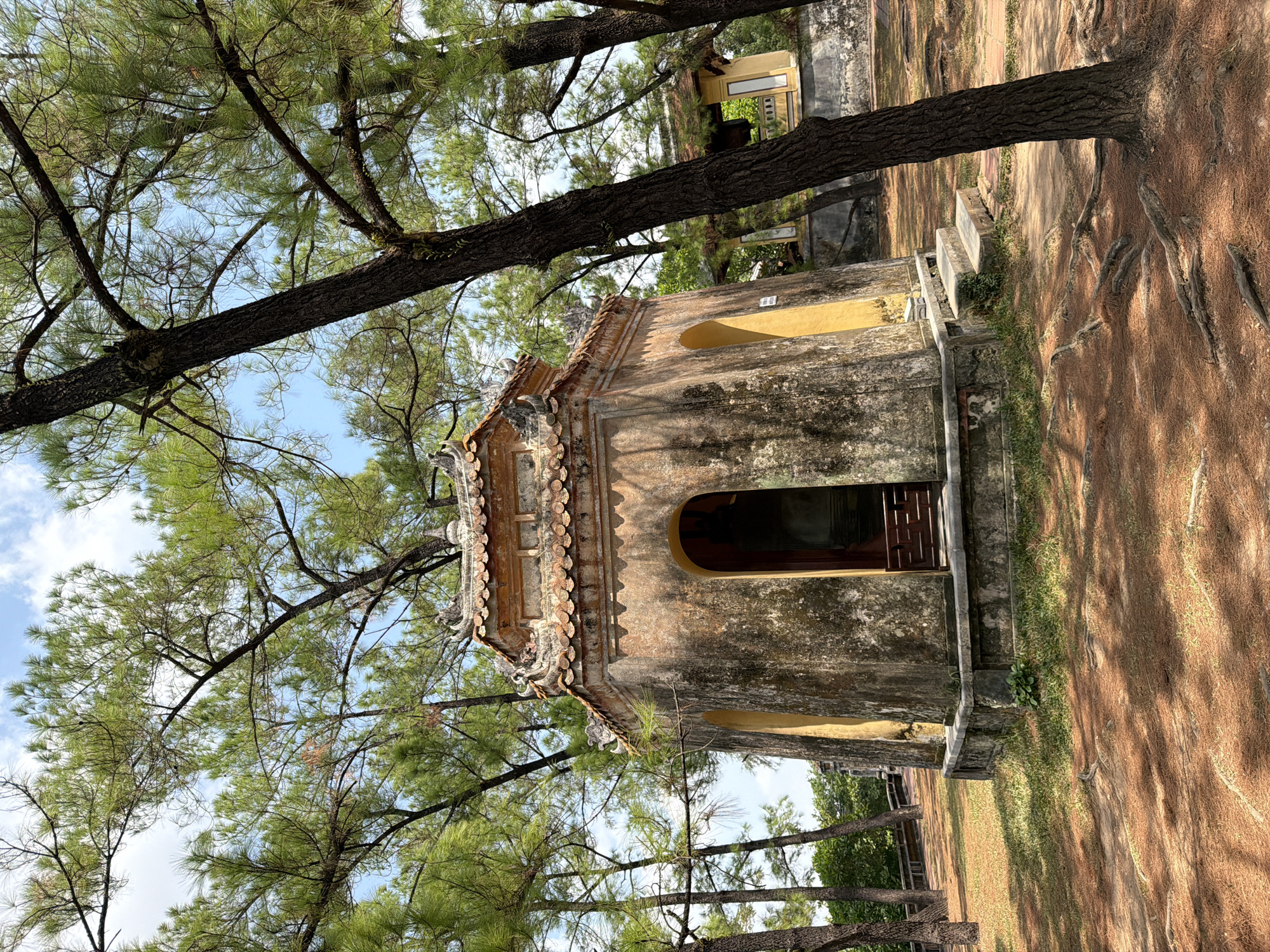 The image captures an octagonal, weathered stone structure with a tiled roof, nestled amongst tall pine trees. The building, with its aged, mottled facade and arched entrance, appears to be a temple or tomb. The entrance is partially open, revealing a dark interior, possibly housing a statue. Yellow-painted alcoves flank the main entrance. Steps lead up to the building, and the ground is covered in pine needles and sparse grass. In the background, another smaller yellow building with a tiled roof is visible. The scene is bathed in sunlight, casting dappled shadows from the trees. The overall impression is one of historical significance and serene isolation. This structure is located in Kim Long Ward, Vietnam, a region known for its historical imperial tombs and temples.