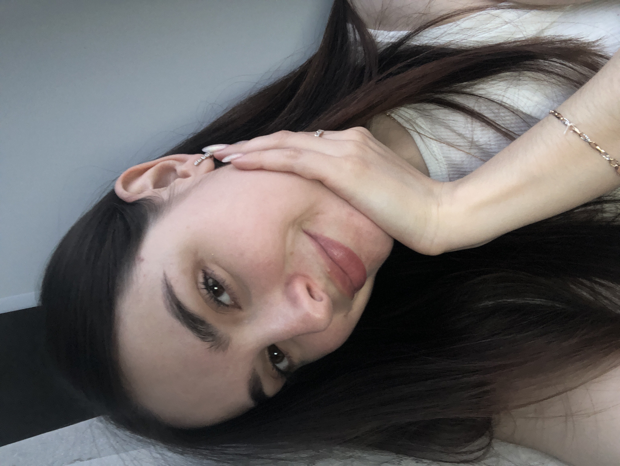 A woman with dark hair is positioned lying down with her head tilted, facing the camera. She is wearing a white tank top. Her right hand is against her cheek. She has a ring on her hand, an earring in her ear, and a bracelet on her wrist. The background is a grey wall and a dark floor.