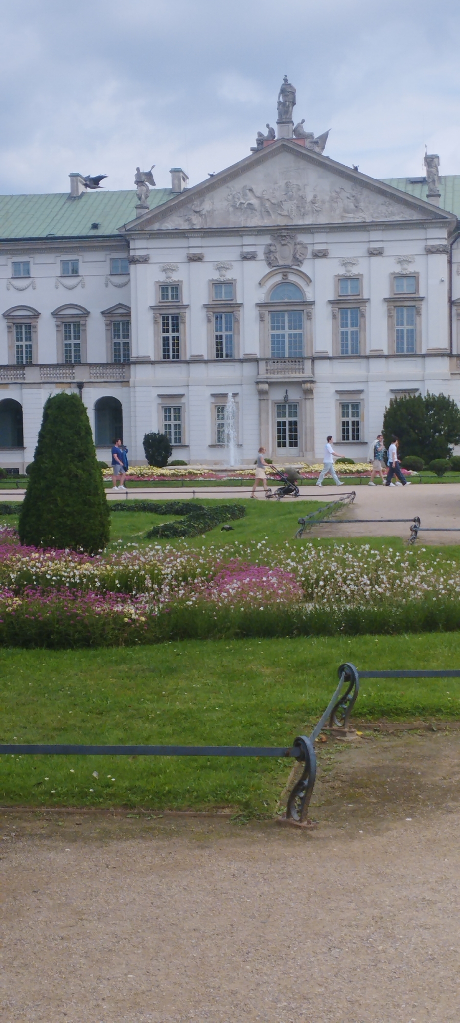 The image shows the Royal Castle in Warsaw, Poland. The castle is a large, white building with a green roof. There are several people walking in front of the castle, and a fountain is spraying water in the background. There is also a large tree on the left side of the image, and a flower bed in front of the castle. The sky is cloudy.