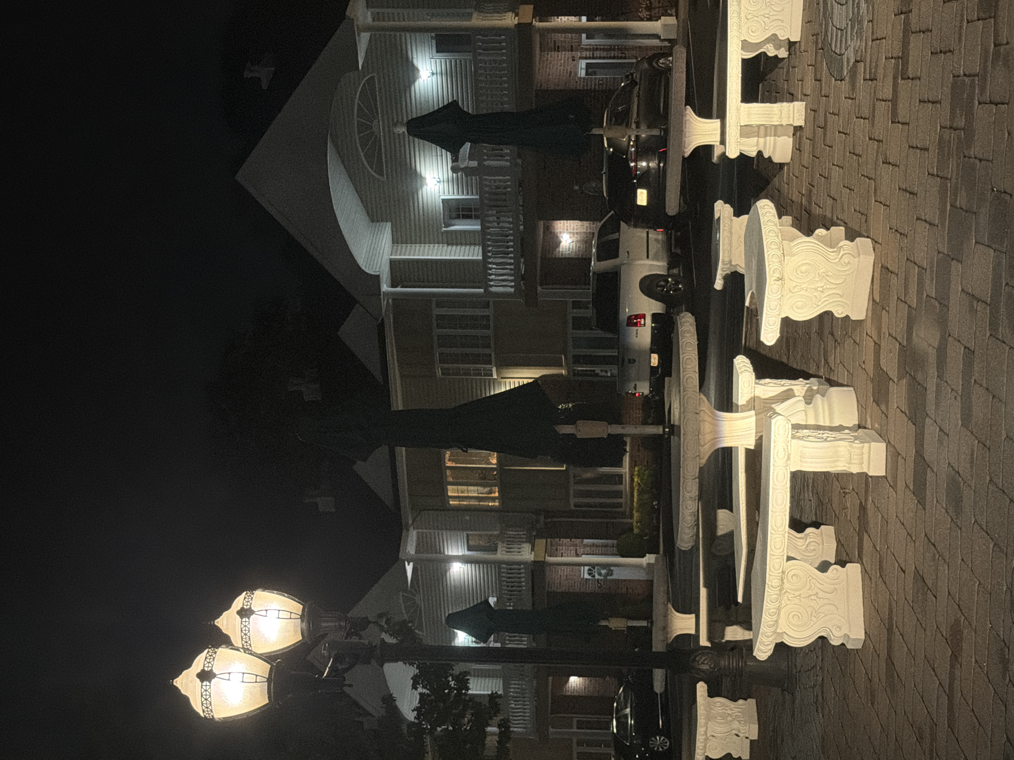 The image shows a nighttime scene in the Village of Amityville, United States of America. In the foreground, there are several ornate white stone benches and tables arranged on a brick-paved area. Two tall, decorative lampposts with illuminated globes stand to the left, casting a warm light. In the middle ground, two large, dark green umbrella-like structures are visible, likely protecting the seating areas. Beyond these, a row of buildings with balconies and illuminated windows suggests residential or commercial units. Several vehicles, including a white pickup truck and a dark sedan, are parked in front of the buildings. The sky is dark, indicating it is nighttime.