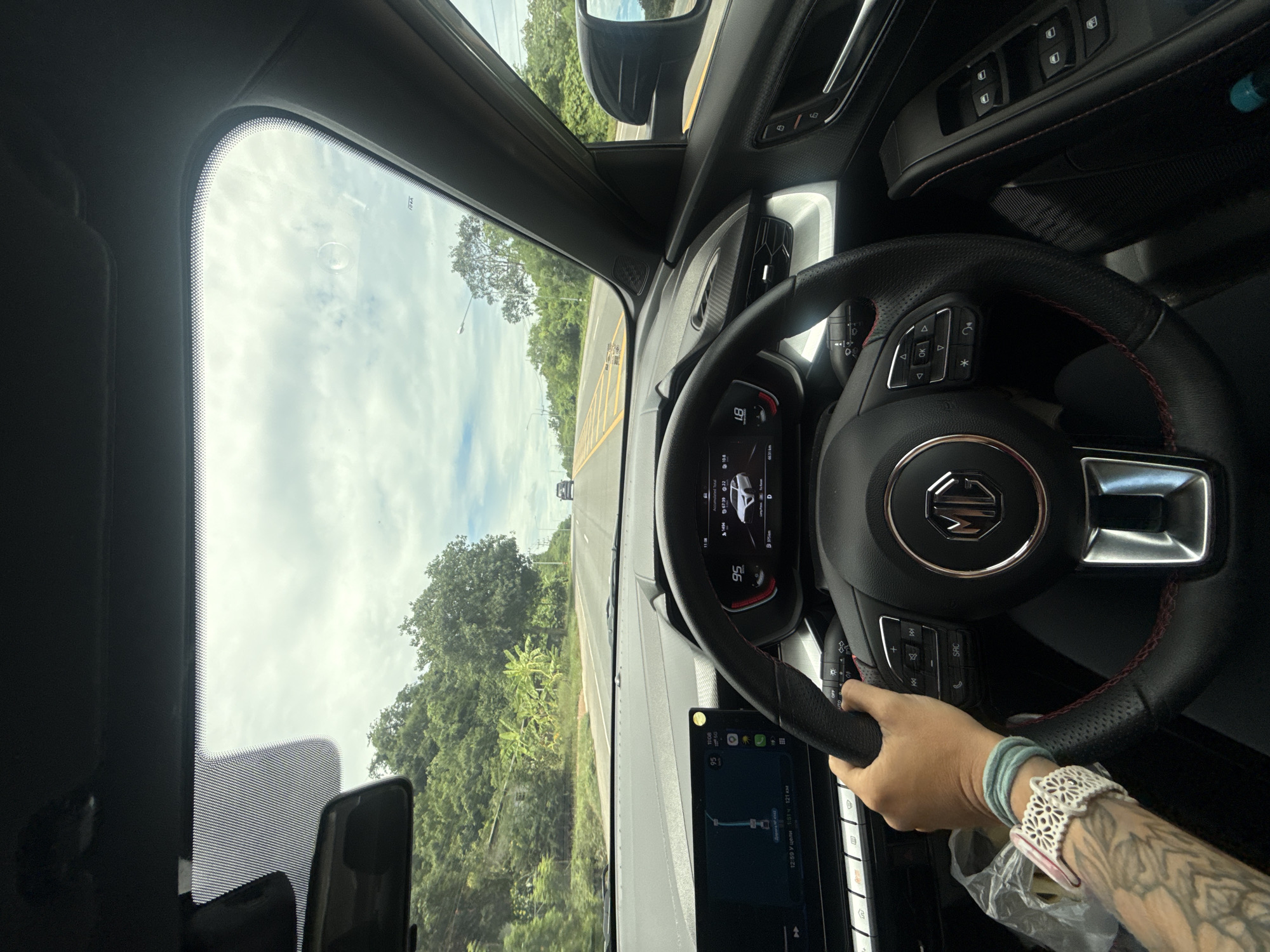 The image shows the interior of a car, viewed from the driver's perspective, as it travels along a two-lane road. The driver's left hand, adorned with tattoos and bracelets, is on the steering wheel. The dashboard features a digital display showing speed as 95 km/h and other vehicle information, as well as a central touchscreen with navigation. Outside the car, the road is lined with trees and lush vegetation, indicative of a tropical or subtropical climate. The sky is partly cloudy. The car is traveling on a road in the vicinity of Don Sak, Thailand, a district known for its coastal access and proximity to islands.