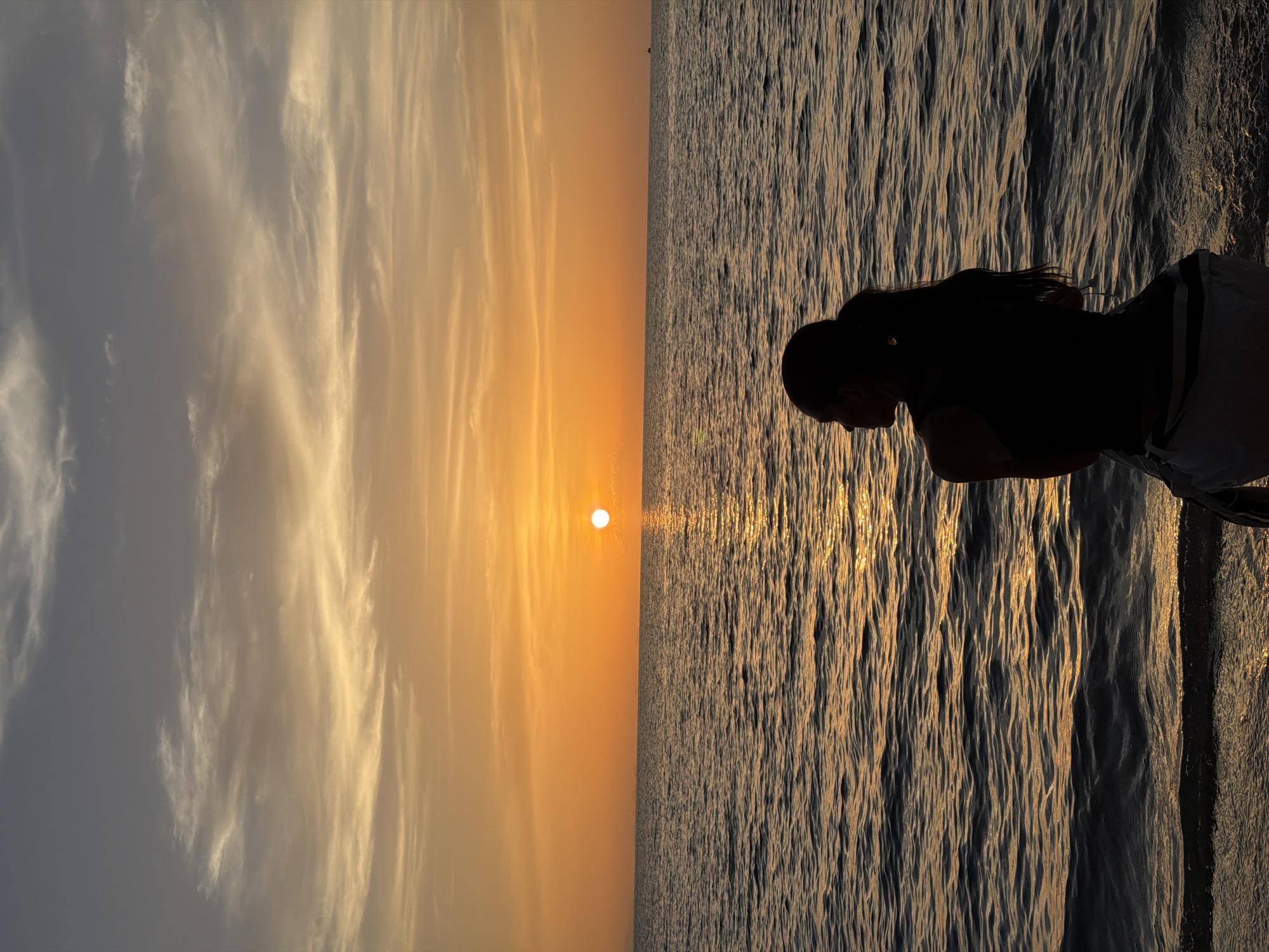 An individual, silhouetted against a vibrant sunset, sits on a beach in Koh Samui District, Thailand. The setting sun, a bright orb of light, casts a warm orange glow across the sky, with scattered clouds tinged with yellow and gray. The ocean stretches out to the horizon, its surface rippling and reflecting the colors of the sunset. The individual is facing away from the camera, looking out at the sea, with their hair pulled back in a ponytail. The foreground shows the sandy shoreline, with gentle waves lapping at the edge. The overall scene evokes a sense of peace and tranquility.