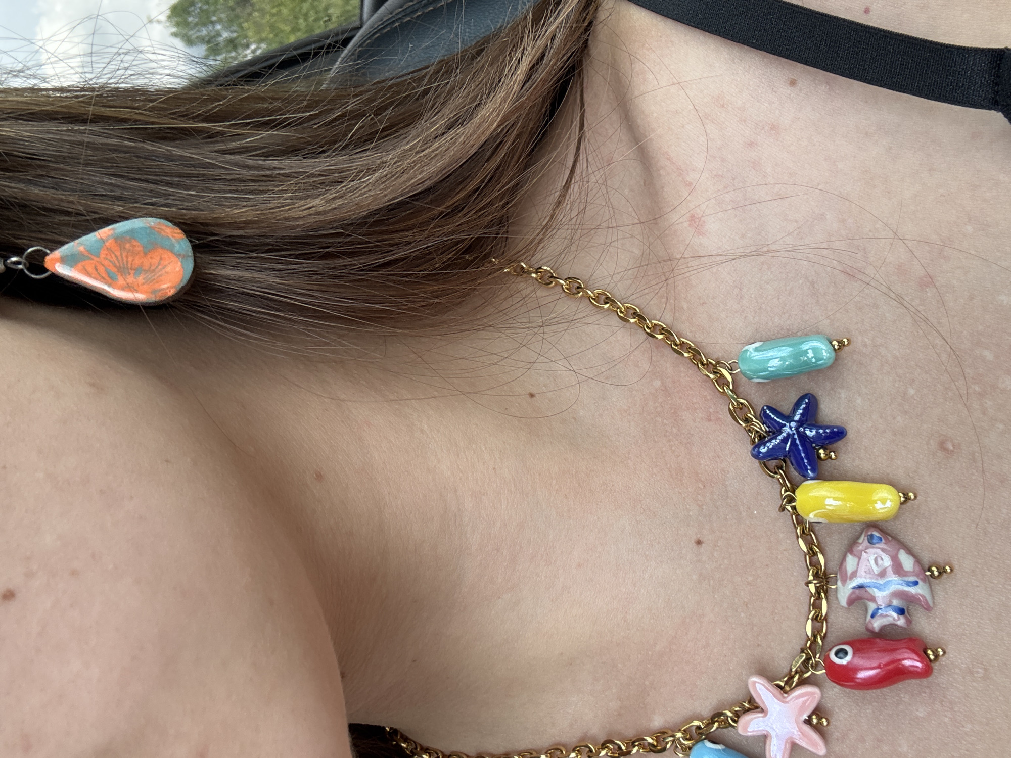 A medium shot shows a person's neck and upper chest area, adorned with a gold chain necklace featuring colorful, nautical-themed pendants (starfish, fish, turtle). A small tear-shaped earring with an orange floral pattern is visible near the person's hair. The person has light skin, with visible moles. A black strap, possibly from clothing, is visible across the upper chest. The photo was likely taken indoors with natural light. The location in the city of San Andrés Cholula, Mexico is not apparent from the visual information.